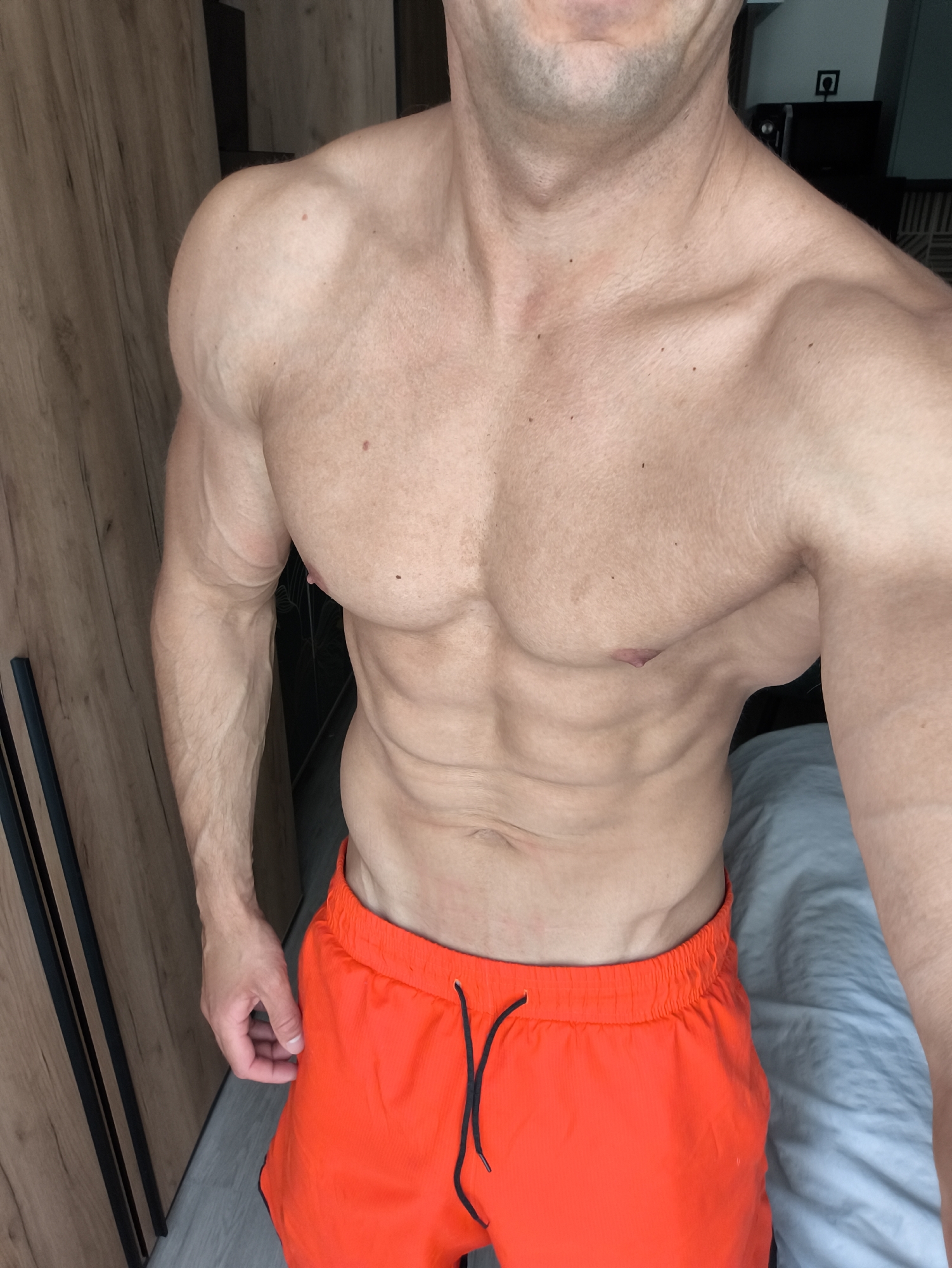The image shows a muscular man with defined abs and pecs wearing orange athletic shorts. He is standing in what appears to be a bedroom. A wooden wardrobe is visible in the background on the left. A bed covered with a light blue sheet is visible on the right. The location is not identifiable from the image. The action depicted is the man posing, showing his physique. No interaction between people is shown.