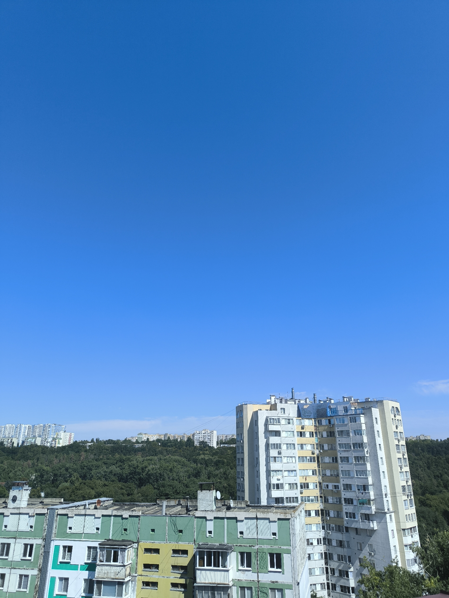 The image shows a view of residential buildings against a clear blue sky. In the foreground, there are several apartment buildings of varying heights and colors, including some painted in green and yellow. Behind these, a densely wooded hill rises, and on top of the hill, more apartment buildings are visible in the distance. The buildings in the foreground appear to be older Soviet-era panel buildings, characteristic of many residential areas in Chișinău, Moldova. The overall scene suggests a typical urban landscape with residential blocks and green spaces.