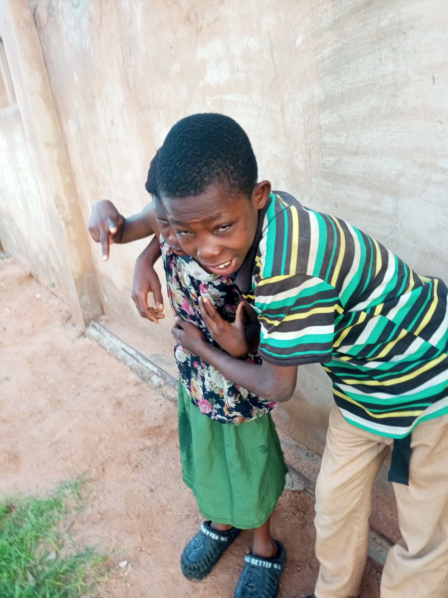 Two children, likely siblings, are standing outdoors next to a textured, beige wall in what appears to be a residential area in Tempure, Nigeria. The child on the left, facing the camera, has their left arm around the other child's shoulders and is pointing downwards with their left index finger. This child wears a floral print blouse and a green skirt, and black sandals. The child on the right is leaning slightly forward, with their right arm around the other child's waist. This child wears a striped t-shirt with green, black, yellow, and white horizontal stripes, and tan pants. The ground is a mix of reddish-brown dirt with patches of green grass. The wall behind them shows some signs of wear and tear.