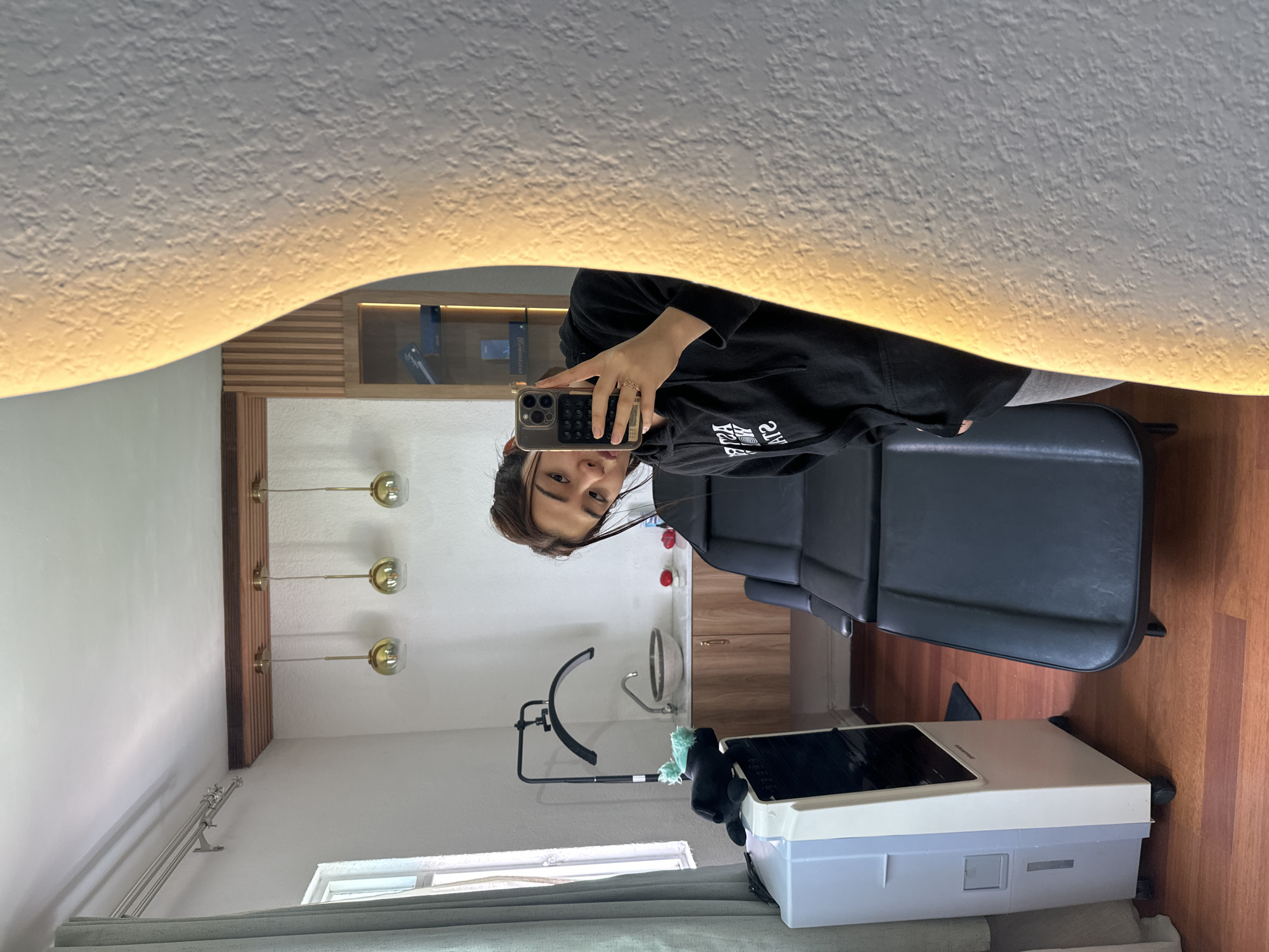 The image shows a selfie taken in a salon, reflected in a large, irregular-shaped mirror with a lighted border. A person is holding a smartphone, partially visible in the mirror, wearing a black hoodie. Behind the person, the salon setting includes a black salon chair, wood-paneled walls with mounted lights, and a white device on wheels positioned near a window. The bottom of the window is covered in a grey curtain. The location is Puebla, Mexico.