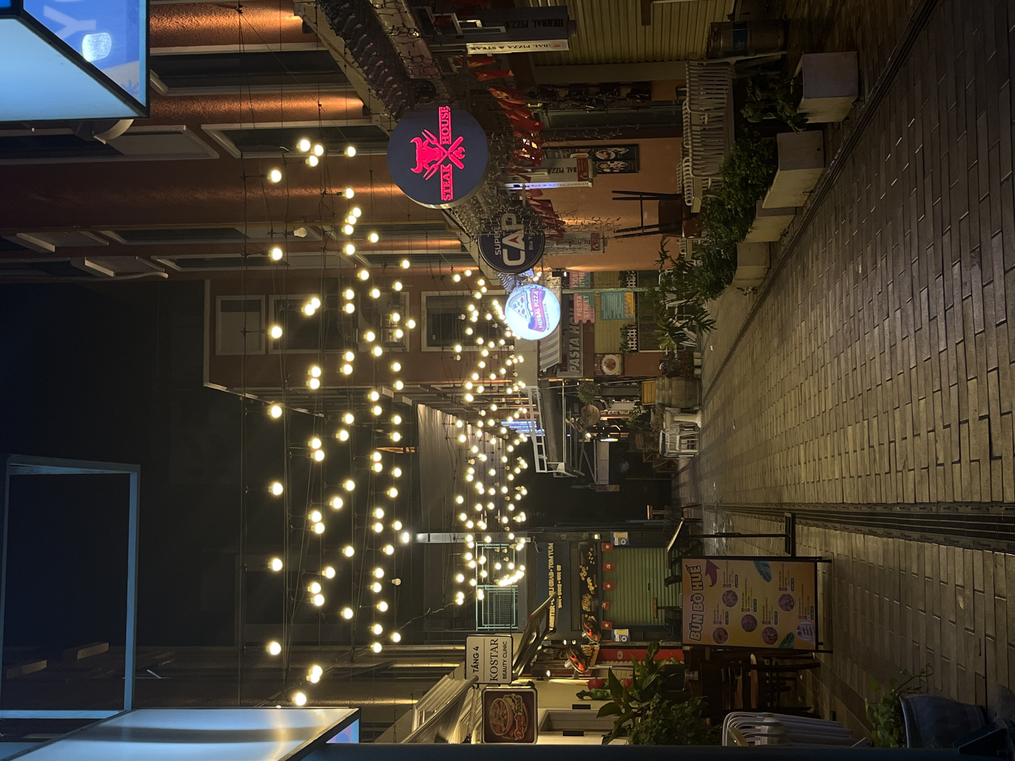 The image shows a narrow pedestrian street at night in Đà Nẵng, Vietnam, lined with restaurants and shops. Strings of illuminated light bulbs hang overhead, creating a warm atmosphere. Restaurant signs are visible, including one for "Steak House," "Super Cap," "Herbal Pizza," and "Bun Bo Hue". Sidewalk seating areas are present, and the street is paved with rectangular paving stones. There is a sign for "Kostar Beauty Clinic". No people are clearly visible in the image.