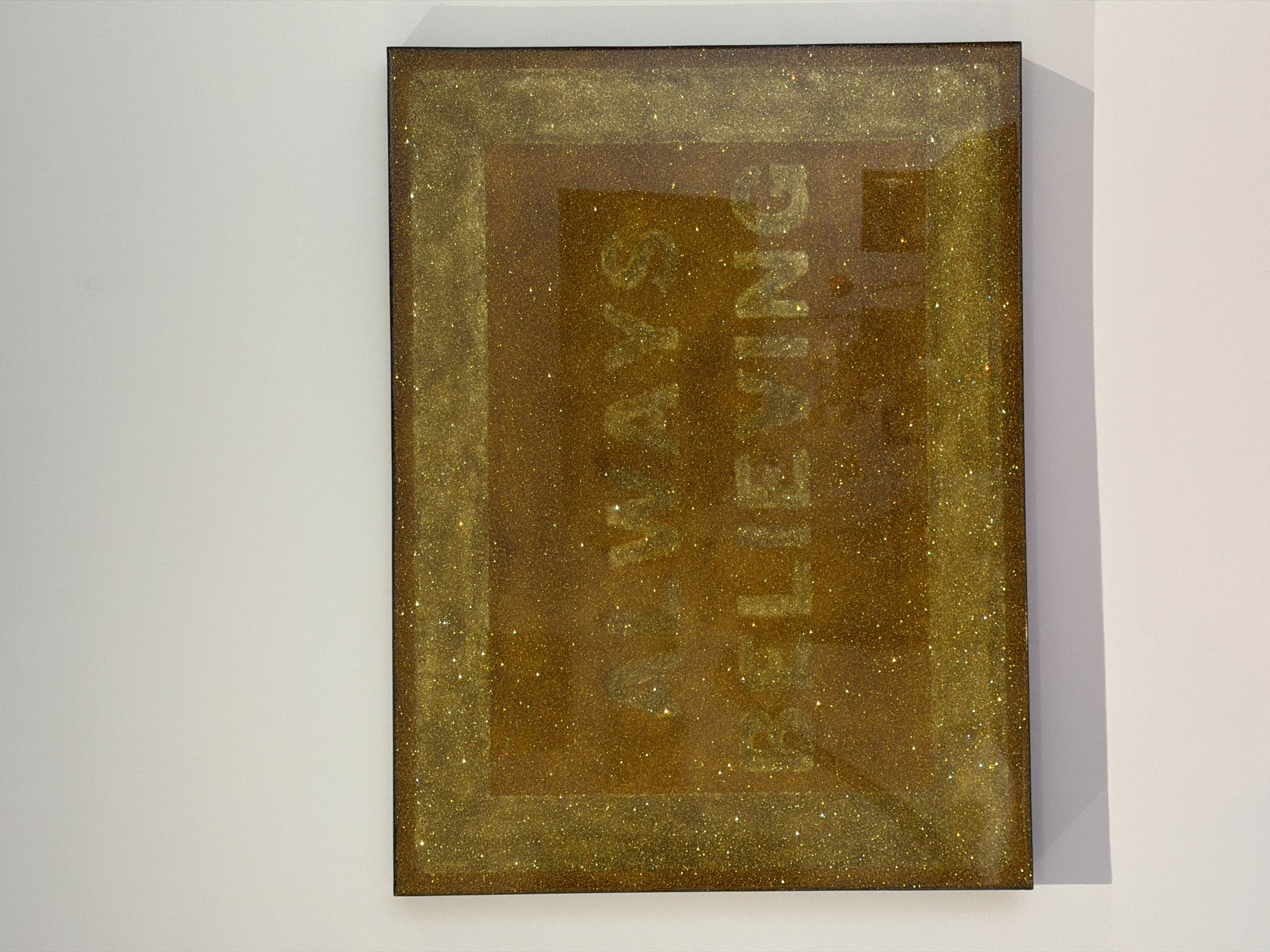 A rectangular gold glitter sign hangs on a white wall. The sign has a frame of solid gold glitter around the edges and a smaller frame of slightly darker glitter inside. Within the inner frame, the words "ALWAYS BELIEVING" are spelled out vertically in gold letters, with each word stacked on top of the other. The sign appears to be indoors, possibly in a home or gallery. The lighting is soft and diffused, creating a warm glow on the sign. No specific location in London can be determined from the image.