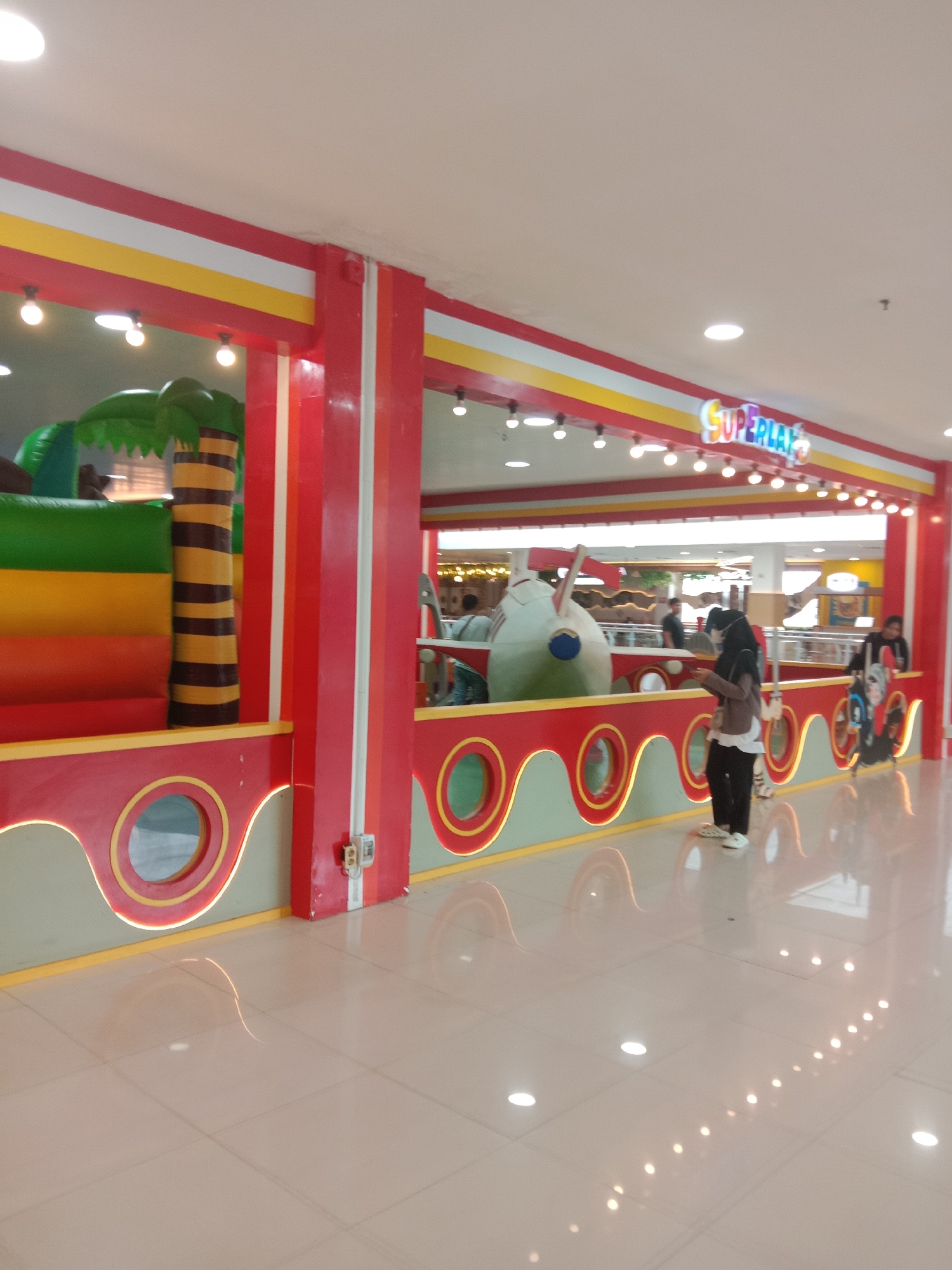 The image depicts an indoor children's play area within a shopping mall, likely in Madiun, Indonesia, identified by the "SUPERPLAY" sign above the entrance. The play area features a vibrant red and yellow color scheme with white accents, and it is illuminated by overhead lighting and decorative string lights. In the foreground, a polished, light-colored tiled floor reflects the surroundings. To the left, a large inflatable structure, resembling a jungle or tropical theme with palm trees and stacked colored sections (green, yellow, orange, and red), is visible. To the right, several people are present. A woman wearing a dark hijab and a light-colored jacket stands looking at her phone. Further to the right, another person is visible with a stroller adorned with cartoon characters. The play area is demarcated by a colorful, wavy barrier with circular openings. In the background, other shoppers and parts of the mall environment can be seen, including a large, white, airplane-like inflatable object within the play zone. The overall impression is one of a recreational space designed for children.