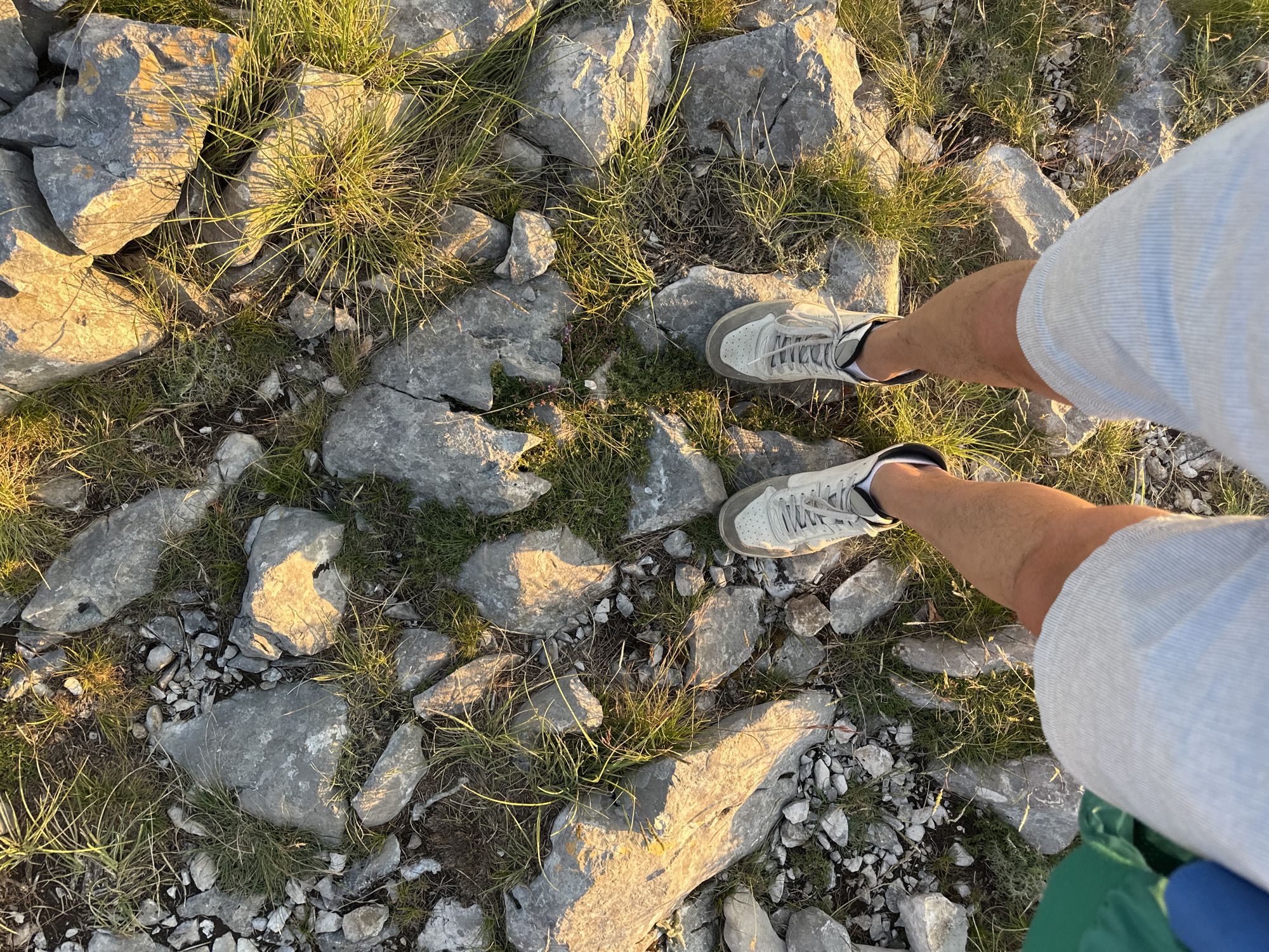 The image shows a first-person perspective of a person's legs and feet standing on a rocky, grassy surface. The person is wearing light-colored shorts and white sneakers. The ground is covered with large, grey stones and patches of dry grass, illuminated by warm, low-angle sunlight, suggesting it is either sunrise or sunset. The location is identified as being in Škaljari, Montenegro. There is a hint of a green object and a blue object on the left side of the frame, partially visible.
