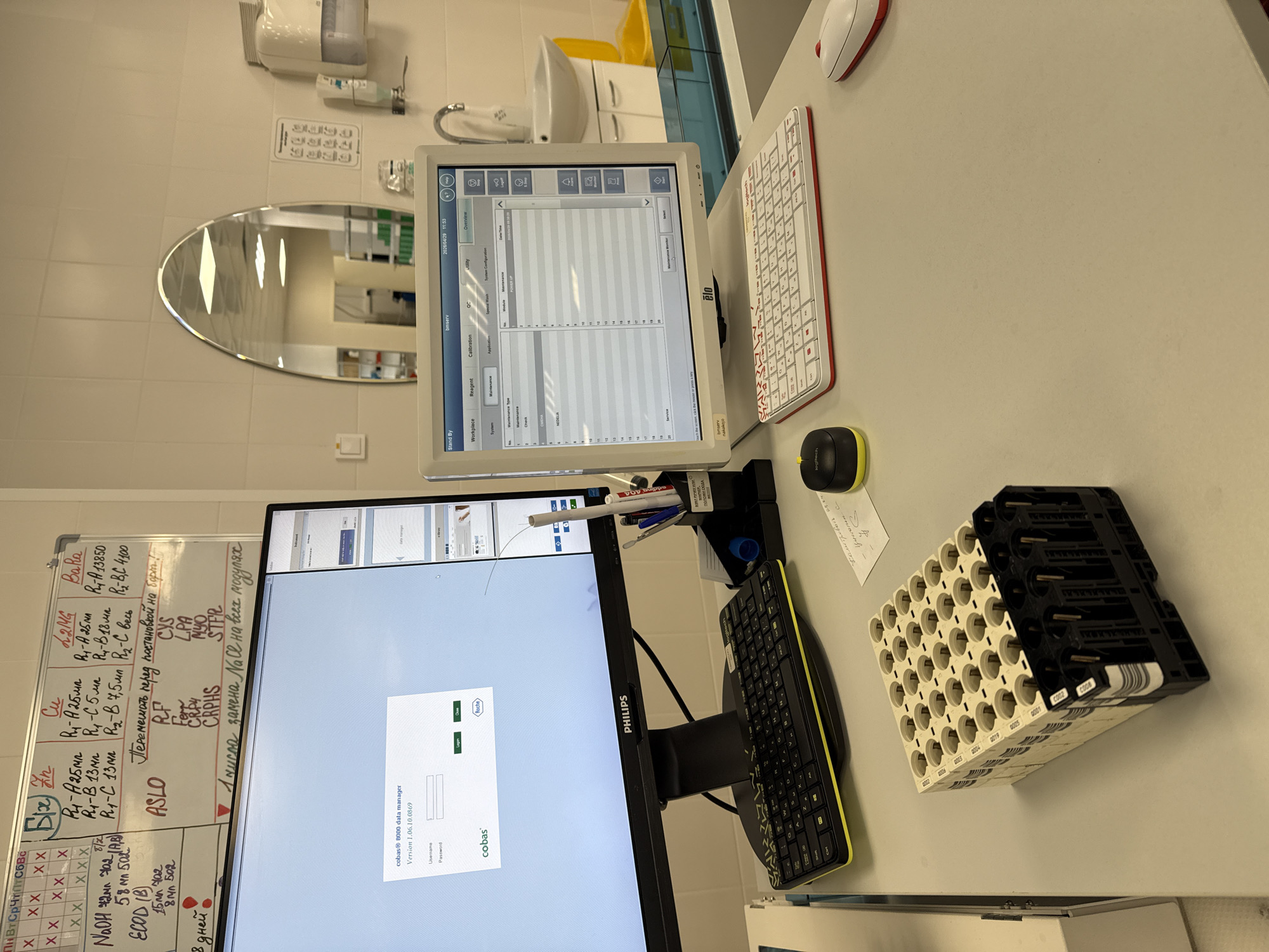 This image captures a desk setup within what appears to be a laboratory or medical testing facility in Moscow, Russia. The scene is devoid of people, presenting a functional and organized workspace.
The main subjects are two computer monitors, two keyboards, and two mice arranged on a light-colored desk. One monitor, a Philips model, displays a login screen for "cobas® 8000 data manager" (Version 1.06.10.0869) by Roche, requesting a Username and Password. The other, an older Elo touchscreen, shows a "Maintenance Monitor" interface with tabs for "Workspace," "Reagent," "Calibration," "QC," "Utility," and "Overview," displaying "2026/04/29 11:53." One keyboard is black (Logitech), the other white and red. A white plastic rack, likely for test tubes or vials, sits on the desk with numbers like "0002" to "C020" visible.
In the background, a white tiled wall features a mirror reflecting parts of the room, including shelving with items. A sink with a faucet, a paper towel dispenser, a soap dispenser, and a yellow container are also visible. A whiteboard mounted on the wall is filled with handwritten notes, predominantly in Russian.
The visible text on the whiteboard includes days of the week ("Пн Вт Ср Чт Пт Сб Вс"), reagent information (e.g., "R₁-A 25мл", "NaOH 2M"), test names ("ASLO", "RF", "Fer", "CRP4", "CYS", "LPA", "MYO", "STFR"), and instructions such as "Перемешать перед постановкой на борт!" (Mix before putting on board!) and "1 числа замена NaCl на всех модулях" (On the 1st, replace NaCl on all modules). The date "2102112" also appears. The monitors' displays, specifically the "cobas" software and the "Maintenance Monitor," indicate that this equipment is used for automated laboratory analysis, possibly for clinical diagnostics. The timestamp on the Elo monitor (2026/04/29 11:53) suggests a future date, which could be a system error or a test environment setting.