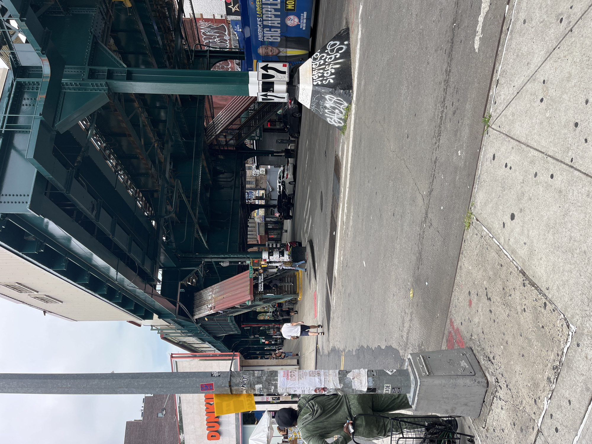 This is a medium shot, rotated image of a street scene in New York City, under an elevated train structure. The train tracks are made of dark green metal beams and supports. The street below is asphalt with white painted lines. A concrete sidewalk runs along the right side of the street. The scene appears to be in an urban environment with storefronts and people present.
In the foreground on the left, a metal pole is visible with stickers and posters attached to it. A person wearing a green hooded sweatshirt and a dark baseball cap is partially visible, pushing a shopping cart. To the right of the pole, a bright orange sign for "Dunkin'" is prominent.
Further down the street, under the elevated structure, several people are walking and standing. Cars are parked and driving on the street. A blue bus with an advertisement for "AMERICA'S GREATEST BIG APPLE" is visible. Traffic signs are mounted on a pole, including directional arrows. Graffiti is visible on the base of the pole with the traffic signs and on the side of the bus.
The overall scene suggests a typical day in an urban area, likely during daylight hours due to the lighting. The weather appears to be partly cloudy. The presence of the elevated train, the mix of commercial signs and graffiti, and the diverse crowd of people are characteristic of New York City. The image captures a slice of daily life in this bustling metropolis.