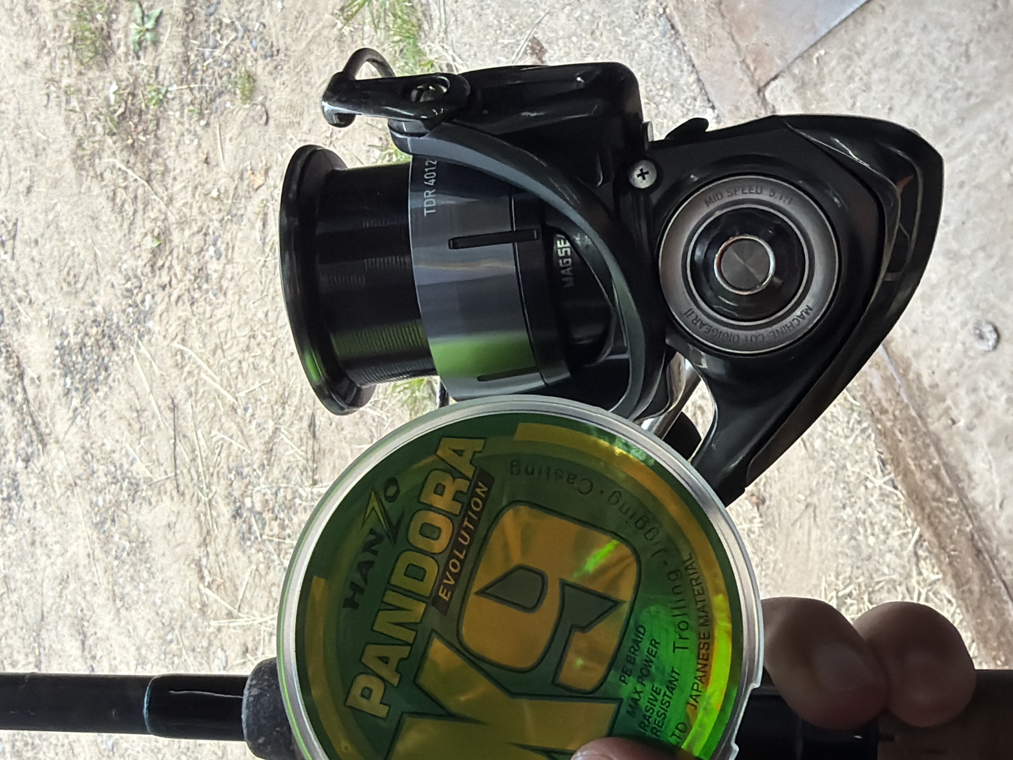 This media file presents a close-up, top-down perspective of fishing equipment held by a person's hand. The main subjects are a black fishing reel and a green and yellow spool of fishing line, positioned over a fishing rod. The background consists of light brown, dry ground, possibly dirt or gravel with sparse vegetation, suggesting an outdoor setting.
The fishing reel is prominently labeled "TDR 4012" and also features the text "MAGSEALED," "MID SPEED 5.1:1," and "MACHINE CUT DIGIGEAR II," indicating its model and specifications. The spool of fishing line, held by the person's fingers at the bottom right, is branded "HANZO PANDORA EVOLUTION." Additional text on the spool describes it as "PE BRAID" with "MAX POWER" and "BRASIVE RESISTANT" properties, made from "JAPANESE MATERIAL," suitable for "Jigging Casting Trolling."
The activity depicted is likely the preparation for or engagement in fishing, with the line being spooled or inspected. The image is well-lit by natural light, indicating daytime and clear weather. The overall scene conveys a rugged, outdoor environment, specifically in Chaikovsky, Russia, implying a fishing expedition or recreational activity.
