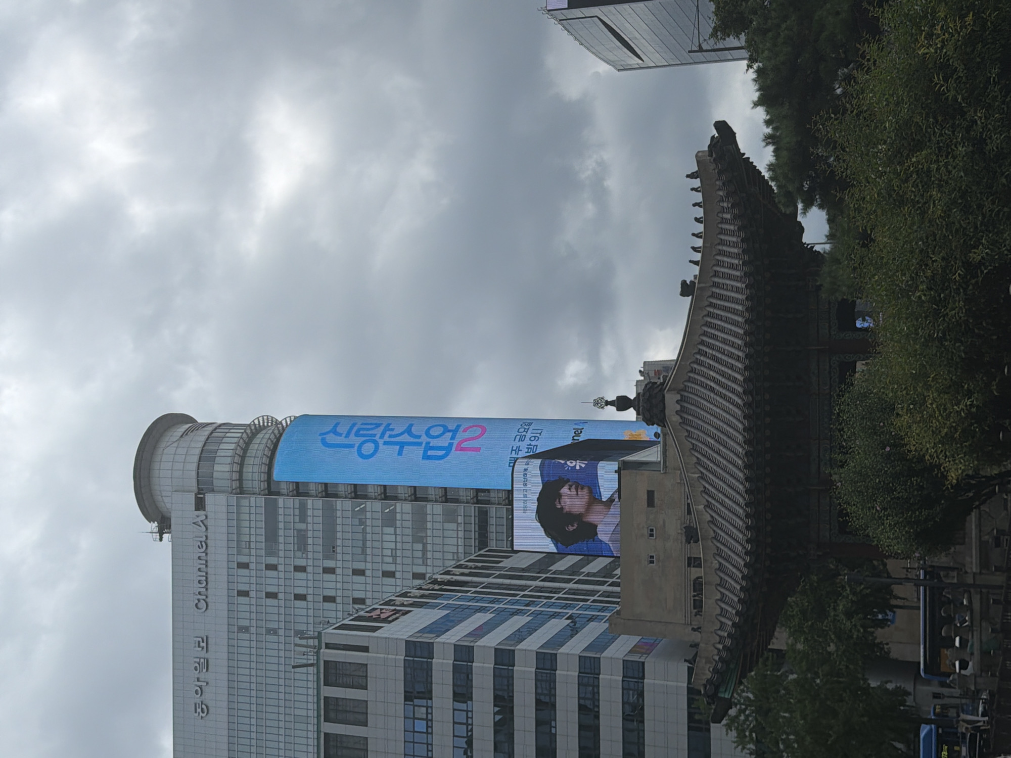 The image shows a view of a city street with a traditional Korean pavilion in the foreground, modern buildings in the background, and a cloudy sky overhead. The pavilion has a dark, ornate roof with intricate carvings and sits amidst lush green trees and bushes.
In the background, a tall, modern building with a curved top is prominent. A large, vertical LED billboard is attached to this building, displaying a blue advertisement with Korean text and a portrait of a young man. To the left of this building is another multi-story building with a glass facade. The overall atmosphere suggests an urban environment in Seoul, South Korea, likely during overcast weather. There are no visible people in the scene, except for a few figures on top of the pavilion, which could be decorative statues or actual people. A bus is also visible on the street below.