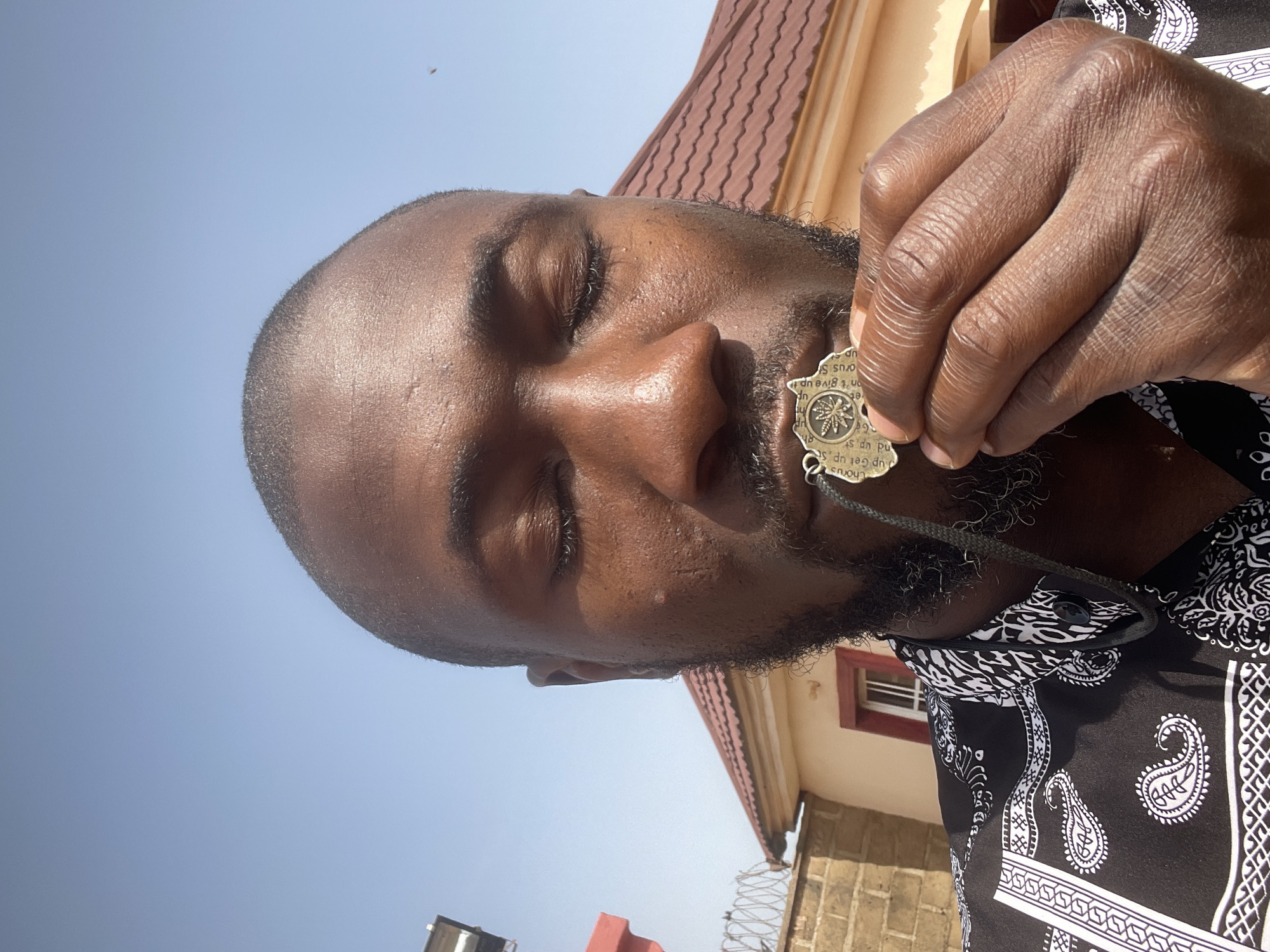 The image shows a close-up of a Black man's face, viewed from the chest up, against a backdrop of a clear blue sky and a house with a red-tiled roof. He has closed eyes and a beard. He is holding a medal or pendant near his mouth with his right hand. The medal appears to have text and an image of a plant on it. He is wearing a black and white patterned shirt. The background suggests a residential area. This scene is likely located in Rayfield, Nigeria, a known residential and commercial area.