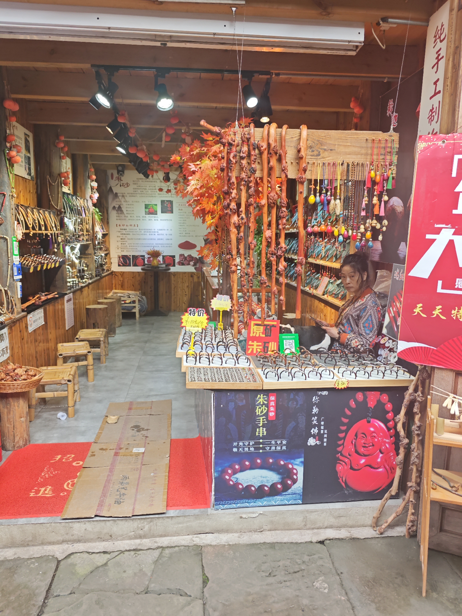 The image depicts the interior of a small shop in Shapingba District, China. A middle-aged Asian woman sits behind a counter displaying numerous pairs of eyeglasses and other small items. She is wearing a colorful patterned top. A black and white dog sits next to her on the counter. The shop is filled with wooden items, jewelry, and other craft goods. Red lanterns and artificial autumn leaves decorate the ceiling. Posters advertising "cinnabar bracelets" and "Maitreya Buddha" hang on the front of the counter, written in Chinese characters. A sign advertising "purely handmade" products is seen on the right. The floor is covered with a red rug and sections of cardboard leading to the entrance.