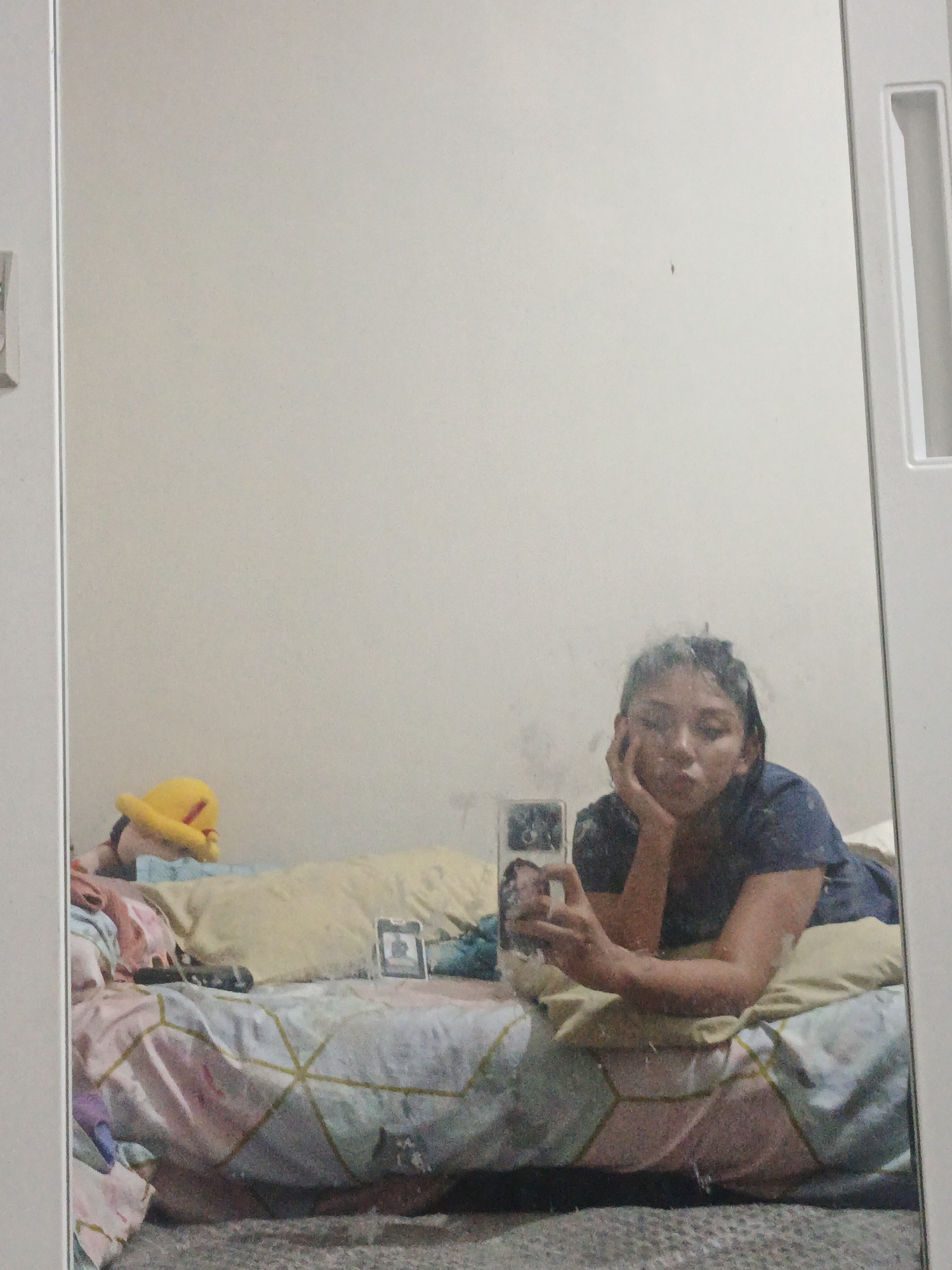 A young woman is pictured lying on a bed in a bedroom. She is facing the mirror, and her reflection is visible. The woman is wearing a dark blue t-shirt and has her right hand supporting her head. Her left hand is holding a smartphone. The bed is covered with a comforter that has a geometric pattern in shades of pink, blue, and yellow. There are several pillows on the bed, one of which is yellow and appears to be a stuffed toy. There is also a small framed picture on the bed. The background of the room is a plain, light-colored wall. The overall impression is of a casual, personal space.