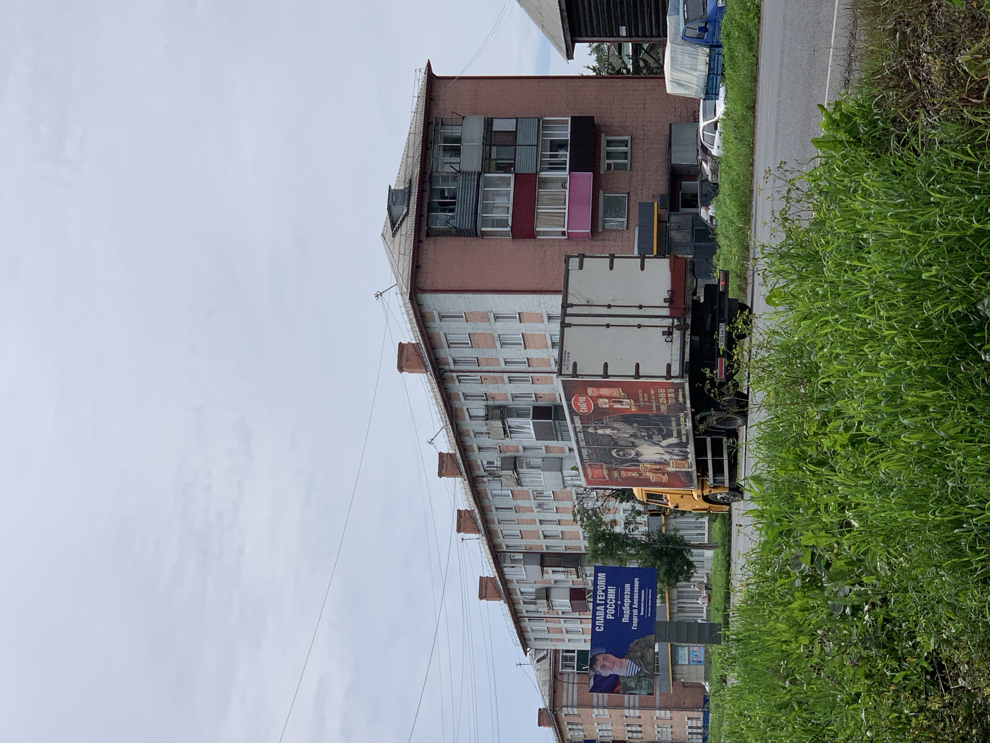 In Prokopyevsk, Russia, a light gray sky with scattered clouds looms over a scene featuring a long, multi-story brick apartment building. The building has a reddish-pink facade with lighter pink vertical stripes and a gray, sloped roof. Multiple balconies with various window and railing configurations are visible, some enclosed, others open.
In the foreground, a paved road runs horizontally, bordered by lush green grass and vegetation. A yellow truck with a white cargo trailer is parked on the side of the road, facing right. The trailer has a large advertisement on its side featuring portraits of people and text in Russian, including "СЛАВА ГЕРОЯМ РОССИИ!" (Glory to the Heroes of Russia!) and a name "Подберезин Георгий Алексеевич" (Podberezin Georgy Alekseevich).
To the left of the truck, a billboard displays a portrait of a man in military-style clothing and similar Russian text. Further down the road, several cars are parked, including a white sedan and a blue truck with a white tarp-covered bed. A darker, wooden building is partially visible on the far right. The overall environment suggests a typical urban or suburban setting in Russia.