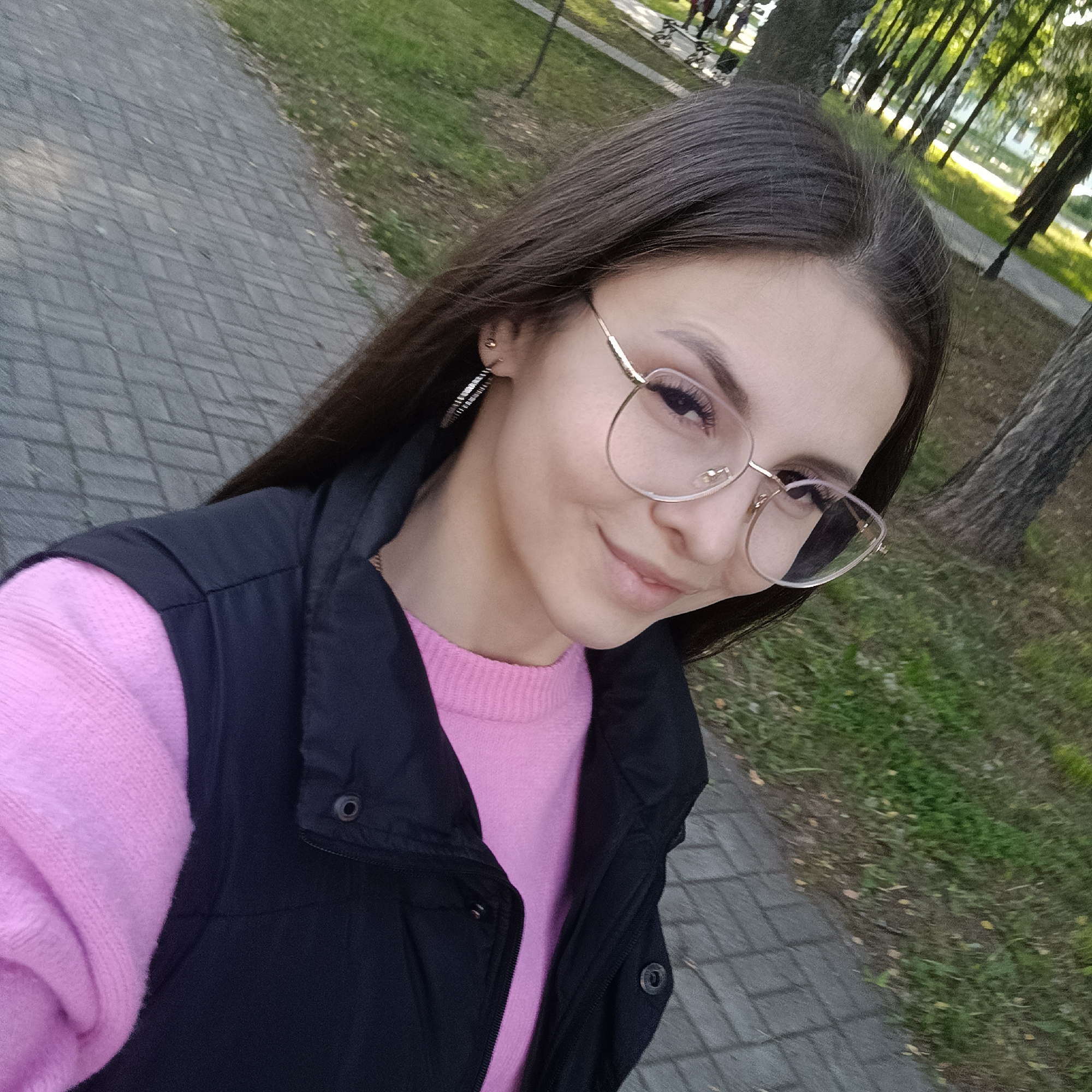 A young woman stands on a brick-paved walkway, with a grassy area dotted with fallen leaves to her right. Behind her are trees, and a building can be seen in the distance. She is wearing glasses, a pink sweater, and a black vest. She faces the camera and smiles.