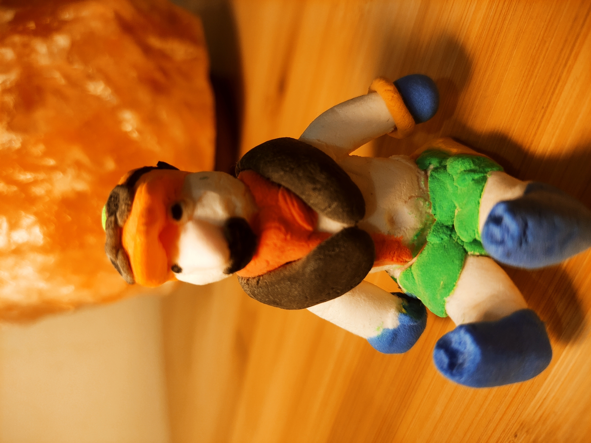 A homemade clay figure rests on a wooden surface, likely a table. The figure, possibly a clown or whimsical creature, has a white body with green, textured accents around the midsection, blue hands and feet, a black and orange head, and dark brown shoulder pads. An orange amorphous object, possibly also made of clay or a different type of material, is to the left of the figure. The scene appears to be indoors, illuminated by a warm light source. The city and specific location are unknown.