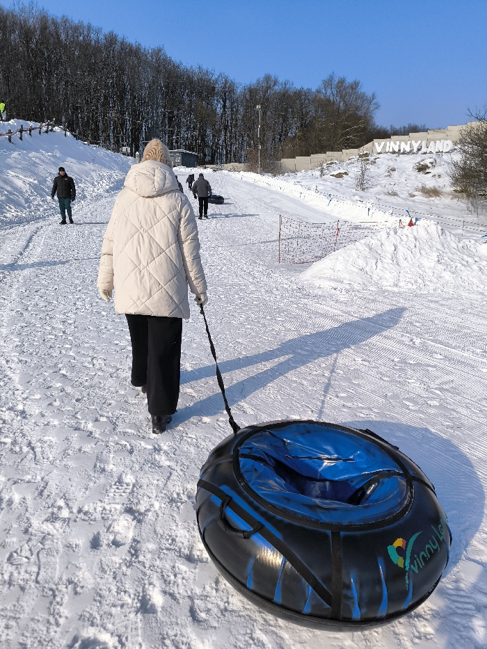 This image depicts a winter scene at Vinny Land in Prybuzke, Ukraine. The foreground shows an individual from behind, wearing a beige quilted jacket, a light-colored knitted beanie, black pants, and dark boots, walking uphill on a snow-covered slope. This person is pulling a black and blue inflatable snow tube, featuring a "Vinny Land" logo, via a black strap. Long shadows are cast on the snow, indicating a low sun angle.
Further up the slope, several other individuals are visible; one person on the left wears a dark jacket and green pants, while two others in the distance also walk uphill, one of whom pulls a dark snow tube. The slope is groomed with visible tracks in the snow, suggesting a tubing or sledding path. To the left, wooden fence posts line the path, and a small building and a light pole are discernible on the hillside. To the right, a large white sign spells out "VINNYLAND" against a snow-covered embankment. Sections of snow netting/fencing are also visible on the right side of the slope. The background features a dense line of bare deciduous trees and some evergreen foliage under a clear blue sky.