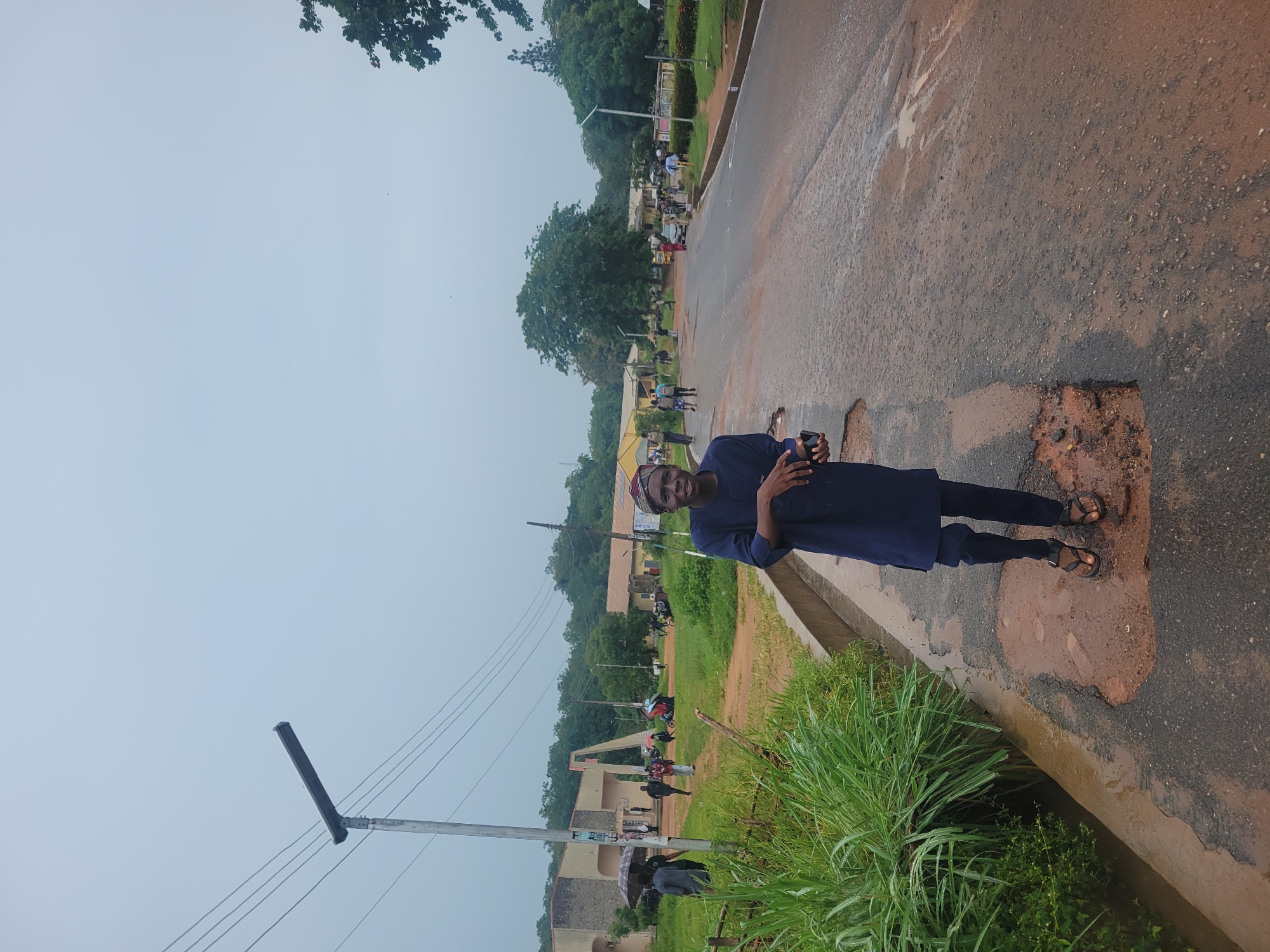 A young Black man, wearing a dark blue tunic and trousers with sandals, stands on a wet, cracked asphalt road, holding a black smartphone in his left hand. He is smiling at the camera. The road is lined with a concrete drainage ditch and overgrown green grass on the left, and on the right, more of the wet road stretches into the distance. In the background, there are several low-rise buildings with light-colored facades, surrounded by lush green trees. A street light with a solar panel is visible on the left side of the image. Several other people, some holding umbrellas, are walking in the distance towards the buildings. The sky is overcast and grey. The scene appears to be an outdoor public space, possibly a campus or a university, given the presence of buildings and students. The overall impression is of a slightly damp and overcast day.