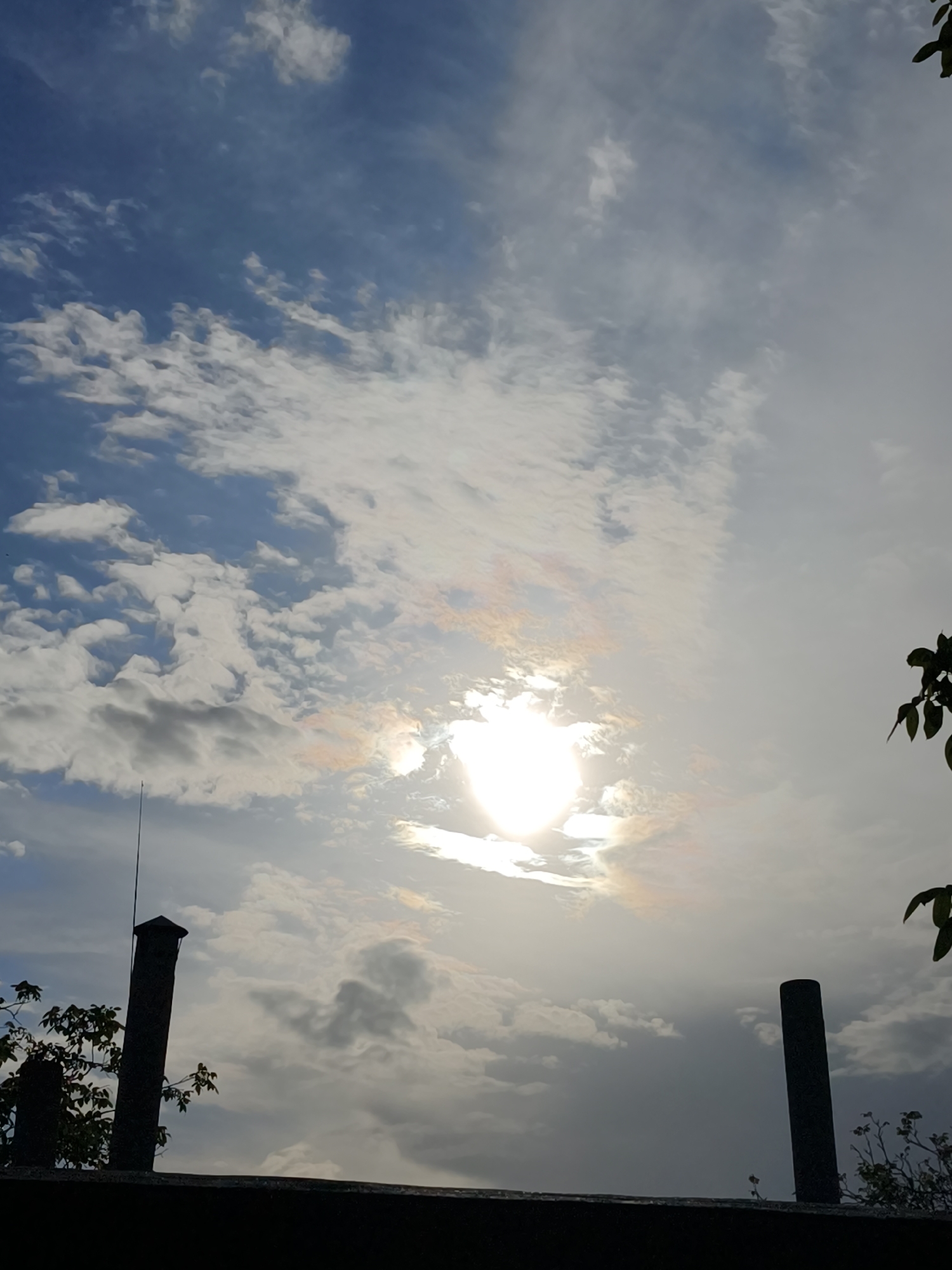 The image shows a view of the sky above what appears to be a building or structure, likely in Kryvyi Rih, Ukraine. The sky features a combination of white and grey clouds against a blue backdrop. The sun is visible, shining brightly through the clouds, with hints of orange and pink hues surrounding it. In the lower portion of the image, there are two cylindrical objects, possibly chimneys or pillars, protruding from a dark, flat surface. Some vegetation, including branches and leaves, is visible on either side of these objects. The foreground is dark, suggesting the perspective is from a lower elevation looking up. The presence of a thin, metallic rod or antenna can be seen extending upwards on the left side of the image.