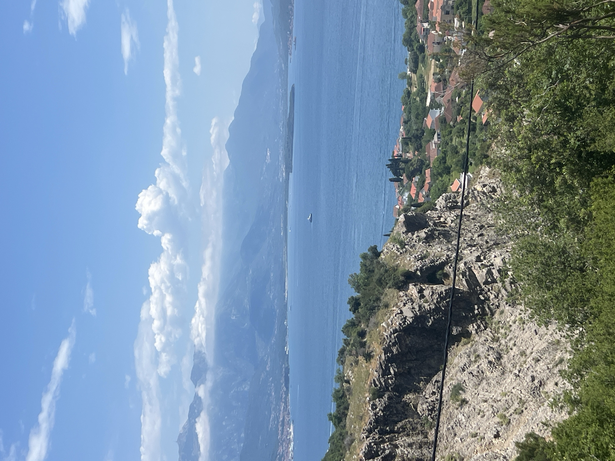 The image captures a scenic view in Baošići, Montenegro. From an elevated vantage point, the turquoise waters of the Bay of Kotor stretch out between mountainous terrain. The sky is partly cloudy with a bright blue hue. On the right, a rocky hillside covered in greenery slopes down to a series of buildings with terracotta roofs, indicating a residential area near the coast. A power line runs diagonally across the right side of the scene. A small boat is visible on the water, implying maritime activity.