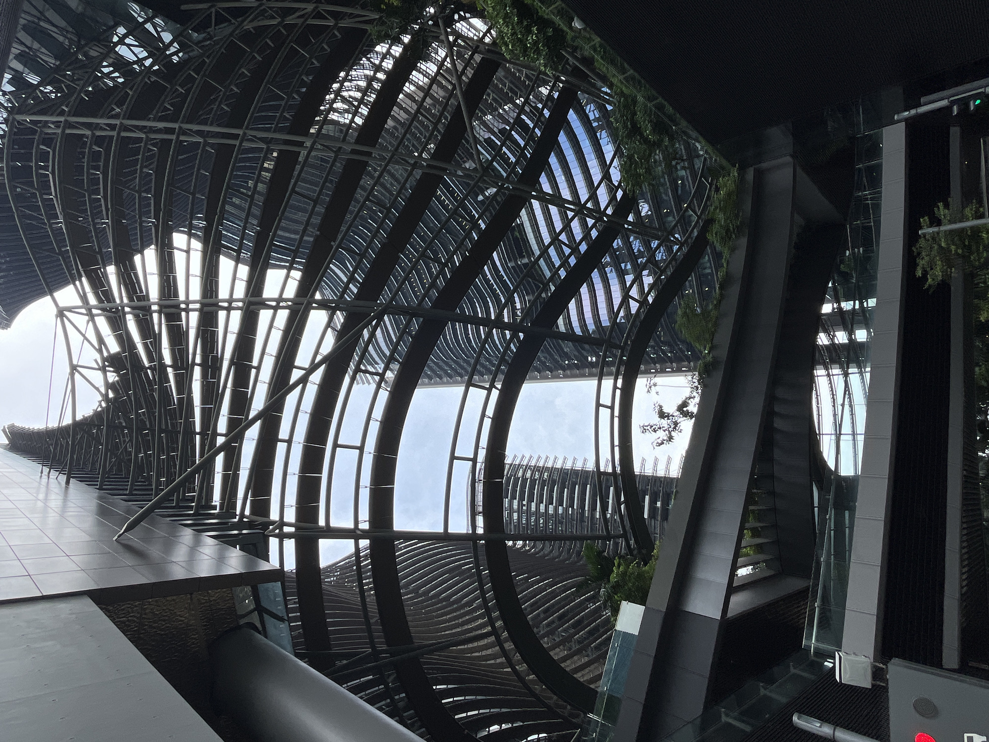 The image depicts a striking architectural design in Singapore. The focal point is a unique, curved metal canopy with a lattice-like structure that arches overhead, creating a dramatic interplay of light and shadow. Beneath this canopy, multiple levels of walkways and bridges are visible, some featuring glass railings and integrated greenery, suggesting a modern, urban park or commercial space. In the background, tall, contemporary buildings with distinctive curved facades and numerous windows rise into the sky. The overall impression is one of a futuristic and sophisticated urban environment, likely a public space designed to blend architecture with nature. The scene is devoid of people.