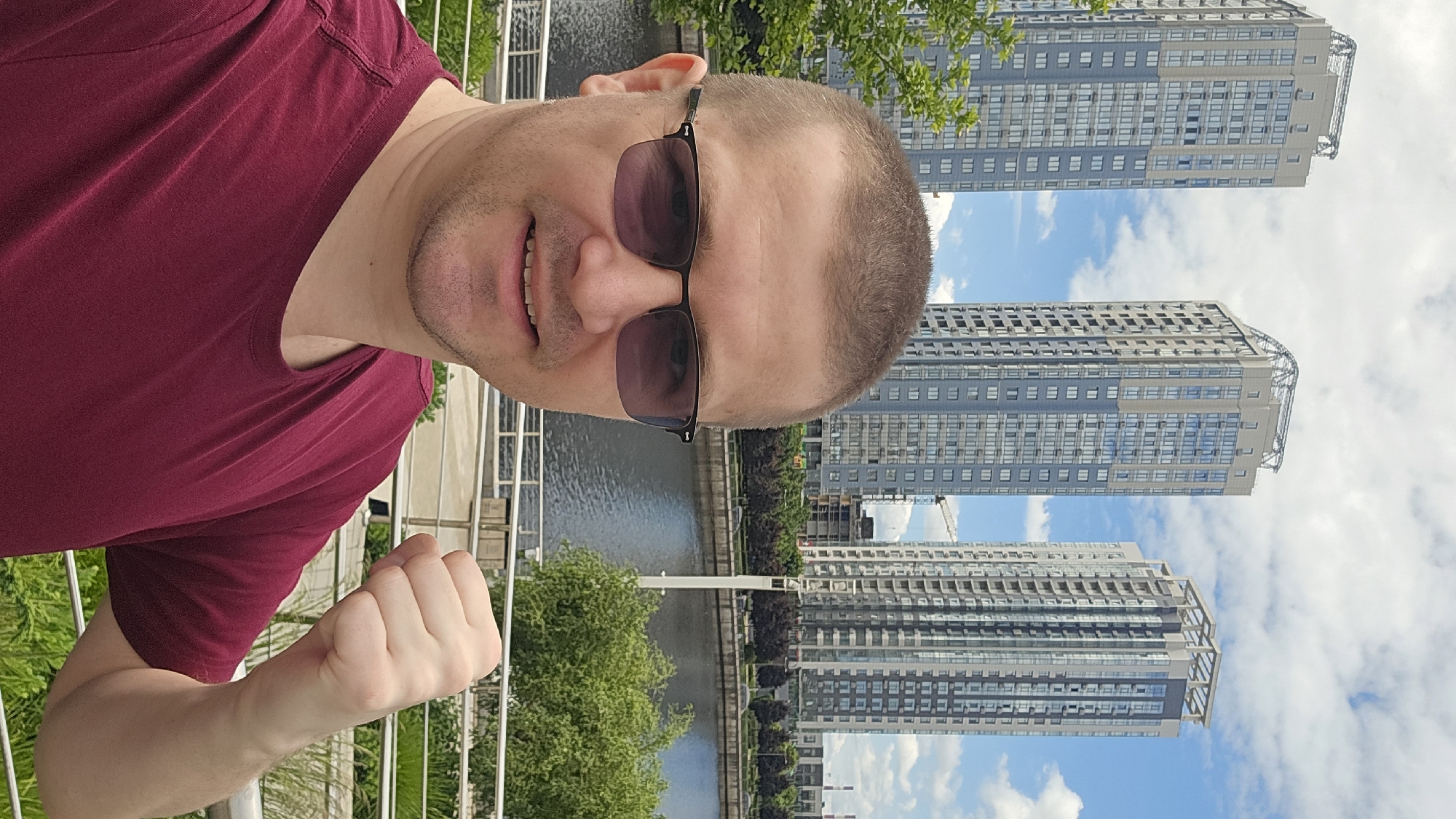 A man in a maroon t-shirt and dark-rimmed glasses is taking a selfie with a slight smile. His right fist is clenched and raised in a celebratory gesture. In the background, three tall residential buildings with grey and blue facades stand against a cloudy blue sky. A body of water, likely a river, is visible in the mid-ground, with lush green trees and a modern walkway in the foreground. The buildings are characteristic of modern urban development, and one of the buildings appears to be under construction, with a crane visible. This scene is located in Kyiv, Ukraine, likely in the Poshtova Ploscha area.