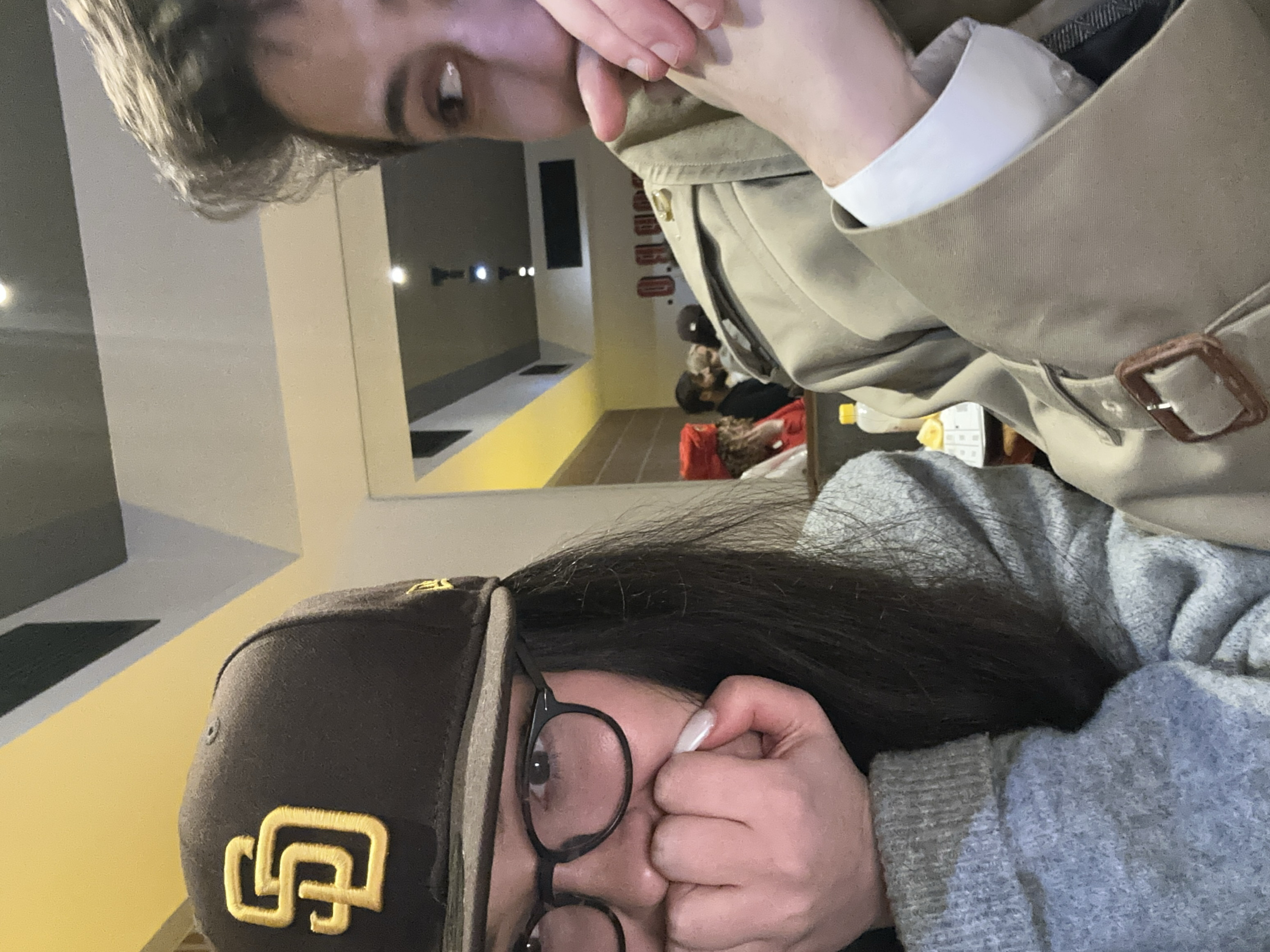 Two people are in the frame. One wears a brown baseball cap with a gold logo, round glasses, and a gray sweater. The other wears a tan trench coat over a white shirt. A mirror in the background reflects an interior hallway with people sitting or lying down. The image is tilted, with the baseball cap wearer appearing in the lower portion of the frame. Both people's faces are partially obscured by their hands.