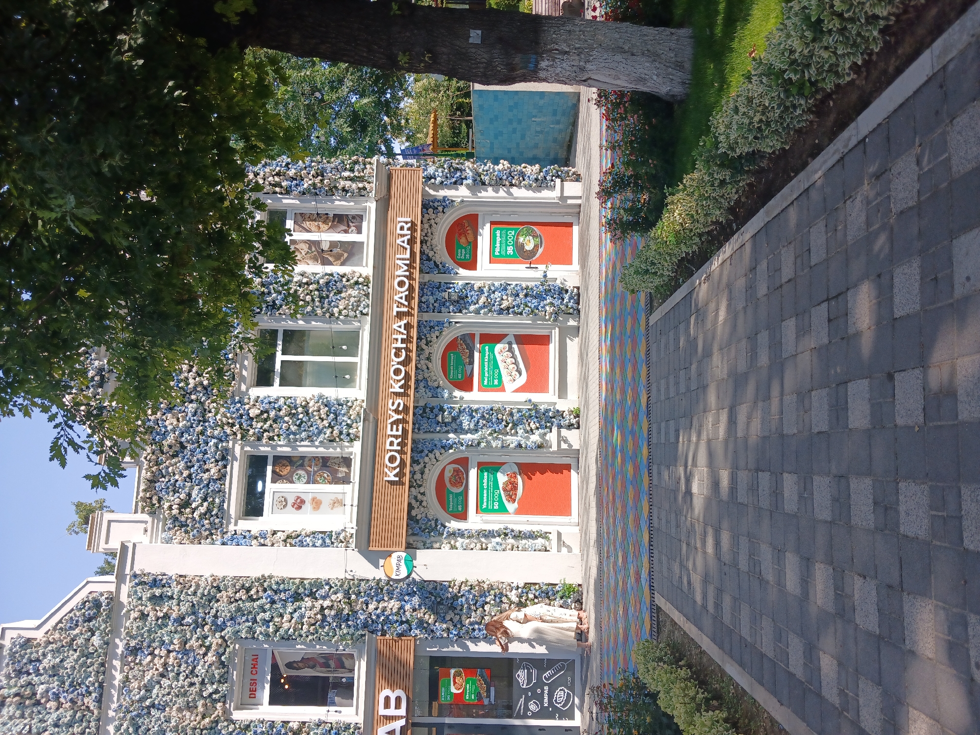 This image depicts a restaurant facade decorated with an abundance of artificial blue and white flowers, creating a charming and eye-catching display. The building has multiple windows and doors, some of which are adorned with promotional posters showcasing food items and prices. Visible text on the building includes "KOREYS KO'CHA TAOMLARI" (Korean Street Food), "DESI CHAI", and "KIMPAB". The posters also list dishes like "Tokpokki", "Yannem chiken", "Mel go'shtil Kimpab", and "Pibimpab" with their respective prices in Uzbek currency.
A woman in a light-colored outfit is seen standing near the entrance, looking down towards the ground. The setting appears to be an outdoor area with a paved walkway leading to the restaurant. The walkway is decorated with a colorful mosaic pattern on one side and grey stone tiles on the other. Green grass and bushes line the walkway and the building. The time of day seems to be daytime, with sunlight casting shadows on the paved area. The overall impression is that of a lively and welcoming establishment, possibly a popular spot for Korean street food in Tashkent, Uzbekistan.
