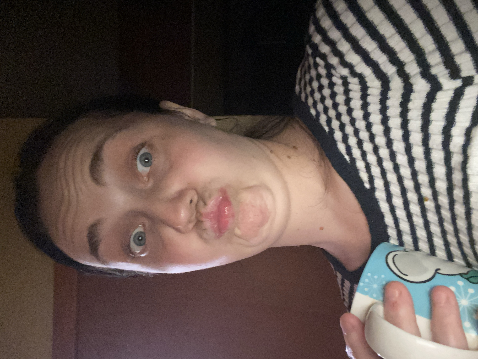 A young woman with light skin and dark hair makes a duck face in a close-up selfie. Her eyes are wide open and blue, and her eyebrows are raised. She is wearing a black and white striped sweater and holding a light blue mug with a white handle. The mug has a cartoonish design with a face and snowflakes. The background is dark and appears to be indoors, possibly a room with a wooden door on the left side. The lighting is dim and casts shadows on her face.