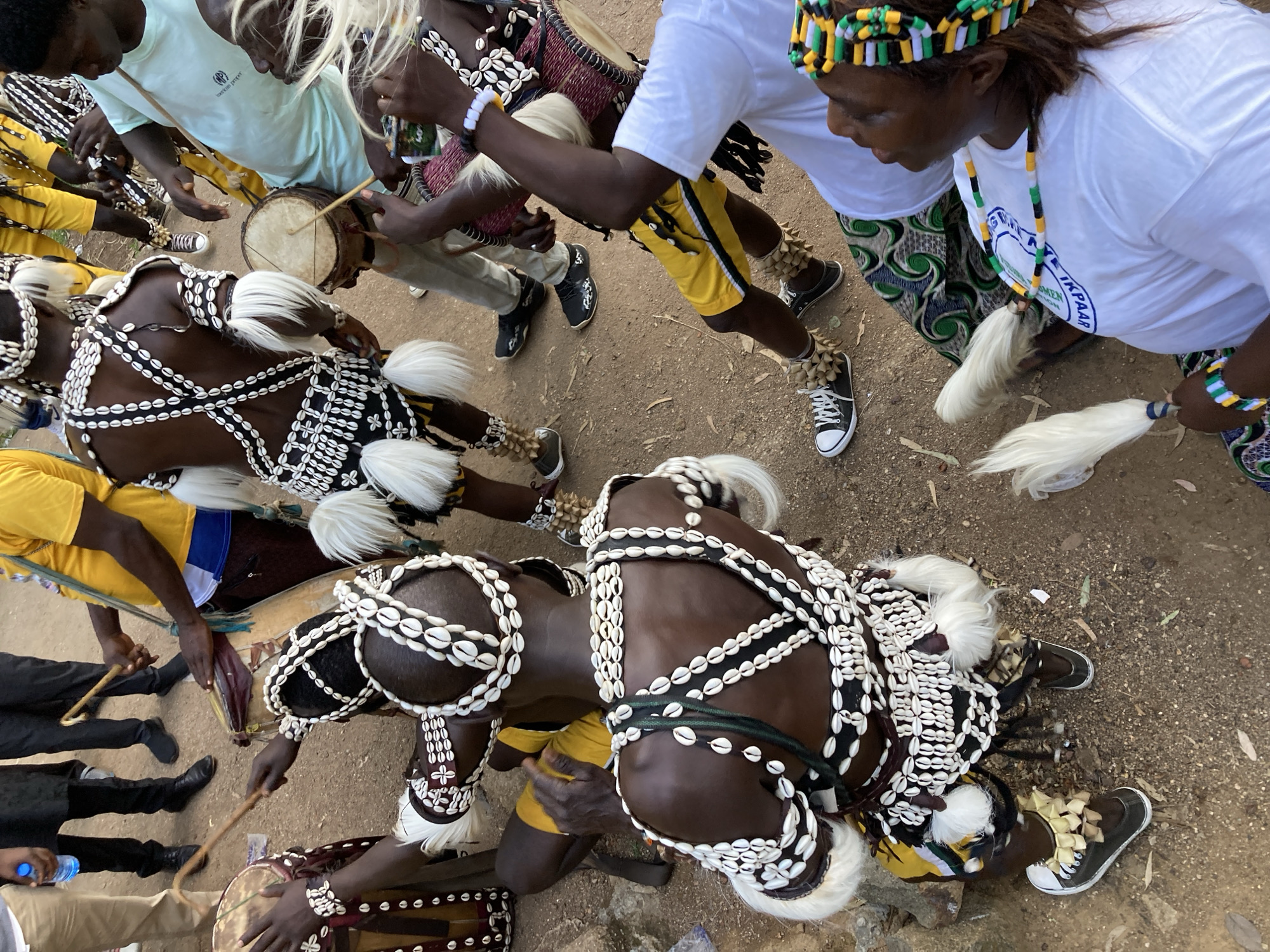 This image depicts a group of individuals engaged in a cultural performance, likely a traditional dance or musical event, on a dusty, outdoor ground that appears to be in the city of Jos, Nigeria. The foreground features two individuals wearing elaborate, traditional attire adorned with numerous white shells and fringe details. One person is seated, playing a small drum with sticks, while the other is standing and also playing a drum. Both are wearing ornate headdresses and ankle decorations made of shells.
In the background, other participants are visible, some also in traditional costumes. One person is wearing a yellow t-shirt and dark trousers, holding a stick. Another is in a light blue t-shirt, and two others are in white t-shirts. One of the individuals in a white t-shirt is wearing a colorful beaded headband and necklace, and is looking upwards. The ground is covered in reddish-brown dirt and some scattered leaves. There are several drums of various sizes present, including a larger, patterned drum held by a person in the mid-ground. The overall atmosphere suggests a lively cultural celebration.