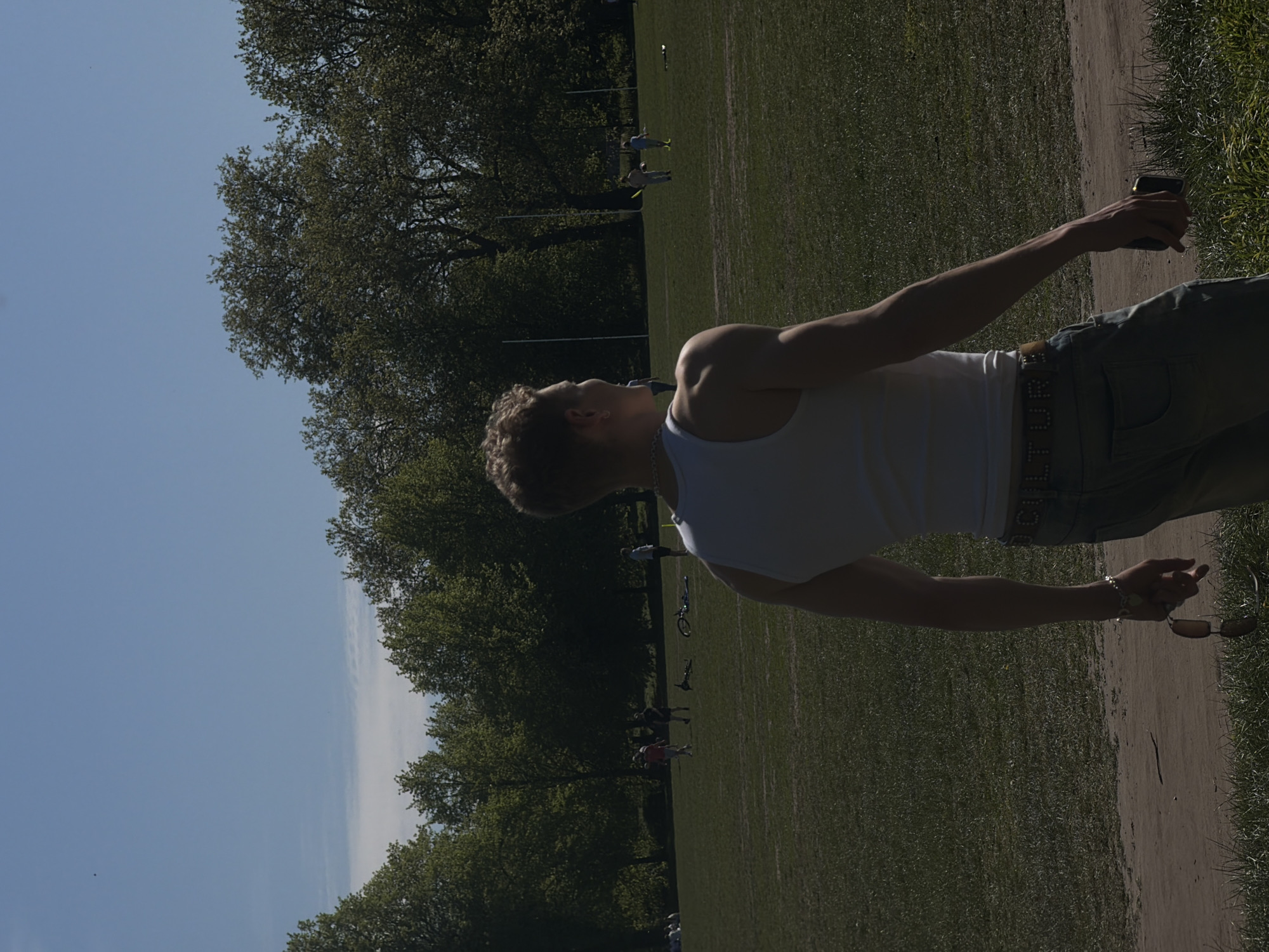 A man with fair hair, wearing a white tank top and light-colored jeans with a belt, stands on a dirt path holding a phone in one hand and sunglasses in the other. He is outside, on a large lawn bordered by trees on one side and the path on the other. Other people are scattered on the lawn in the background, some walking, others biking, and some playing sports. A clear blue sky is visible above the trees. The scene depicts a sunny day in a park environment. Considering the presence of large lawn spaces and trees, the location resembles Hyde Park or Regent's Park in London, United Kingdom.