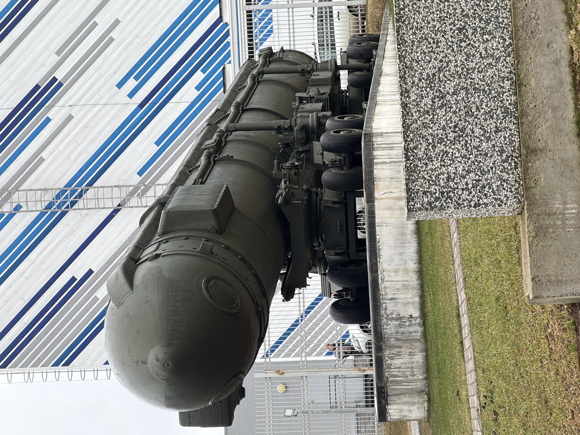 The image displays a large, olive-green ballistic missile mounted horizontally on a multi-wheeled transporter erector launcher (TEL), likely an intercontinental ballistic missile (ICBM). The missile is positioned outdoors, seemingly as a static exhibit or monument.
In the background, behind the missile and its transporter, stands a building or structure with a distinct facade. This facade is predominantly white, adorned with diagonal stripes of varying shades of blue and light grey, creating a modern, abstract pattern. A metal fence or railing, possibly for safety or crowd control, is visible between the missile display and the striped structure.
To the right of the missile, there is a raised platform or wall with a textured surface composed of small black and white pebbles (likely exposed aggregate concrete). Adjacent to this platform and extending further right is a patch of green grass, bordered by a concrete pathway.
A single person is partially visible in the mid-ground, positioned behind the metal fence and partially obscured by the missile's structure. They appear to be observing the exhibit.
The scene takes place during daylight hours, characterized by bright, diffused lighting, suggesting an overcast sky. The overall setting implies a public display, possibly at a museum, a technological exhibition, or as a monument, consistent with the location context of Votkinsk, Russia, a known center for missile production. There is no visible text or identifiable markings on the missile or its transporter.