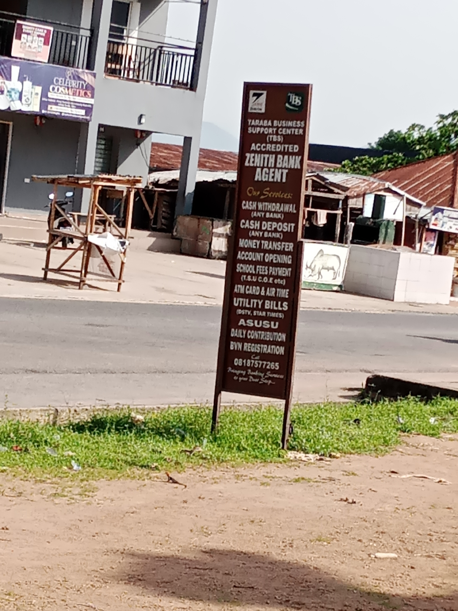 The image shows a street scene in Jalingo, Nigeria. In the foreground, a dark brown sign stands vertically, advertising services offered by the "Taraba Business Support Center (TBS)" which is an "Accredited Zenith Bank Agent". The sign lists various services including cash withdrawal and deposit (for any bank), money transfer, account opening, school fees payment, ATM card and airtime purchase, utility bill payments (DSTV, Star Times), Asusu, daily contribution, and BVN registration. A phone number, "08187577265", is also visible. The sign is positioned on a patch of dry, sandy ground with sparse green grass and some scattered litter.
In the mid-ground, a paved road runs horizontally across the image. Beyond the road, there are several buildings. To the left, a grey building with a balcony and a sign for "Celebrity Cosmetics" is partially visible. In front of this building is a wooden stall with a motorcycle parked behind it. To the right of the Zenith Bank agent sign, there are more structures with corrugated iron roofs, suggesting a market or informal business area.
The scene appears to be in daylight, under a bright sky, though the weather is not explicitly indicated. The overall impression is a typical urban streetscape in a Nigerian city, with commercial activity and various services available. The image is slightly blurred, indicating it was taken with a mobile phone camera from a distance.