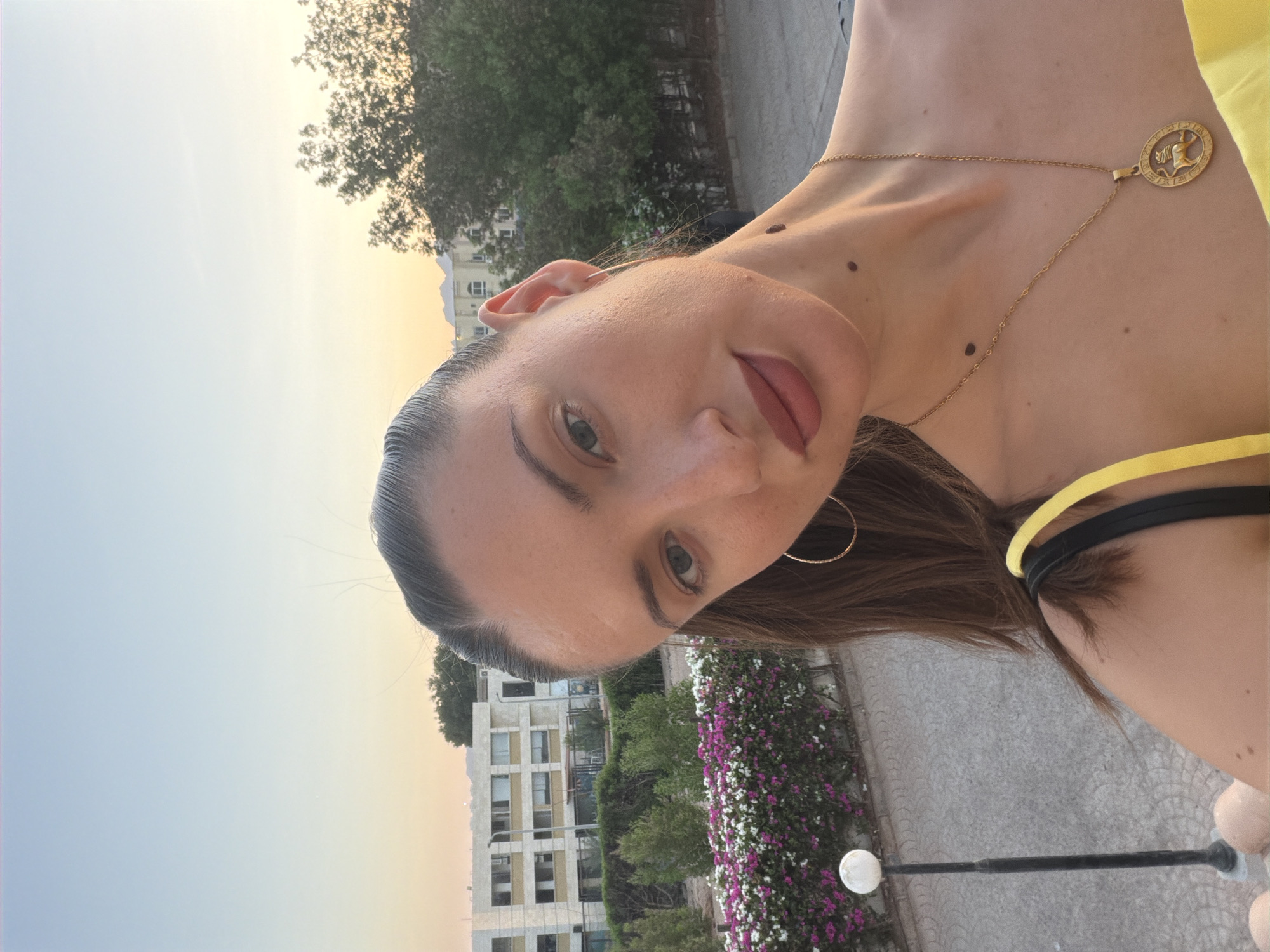 A young woman with long dark hair pulled back in a ponytail takes a selfie outdoors. She is wearing a yellow top, large hoop earrings, and a gold necklace with a circular pendant. She has makeup on her lips and eyes. In the background, there is a building with multiple windows, green trees, and a flowerbed with pink and white flowers. The sky is a clear light blue fading to orange near the horizon, suggesting it is either sunrise or sunset. A lamppost is visible to her left. The overall scene appears to be in a resort or hotel area in Sharm El Sheikh, Egypt, given the architecture and landscaping.