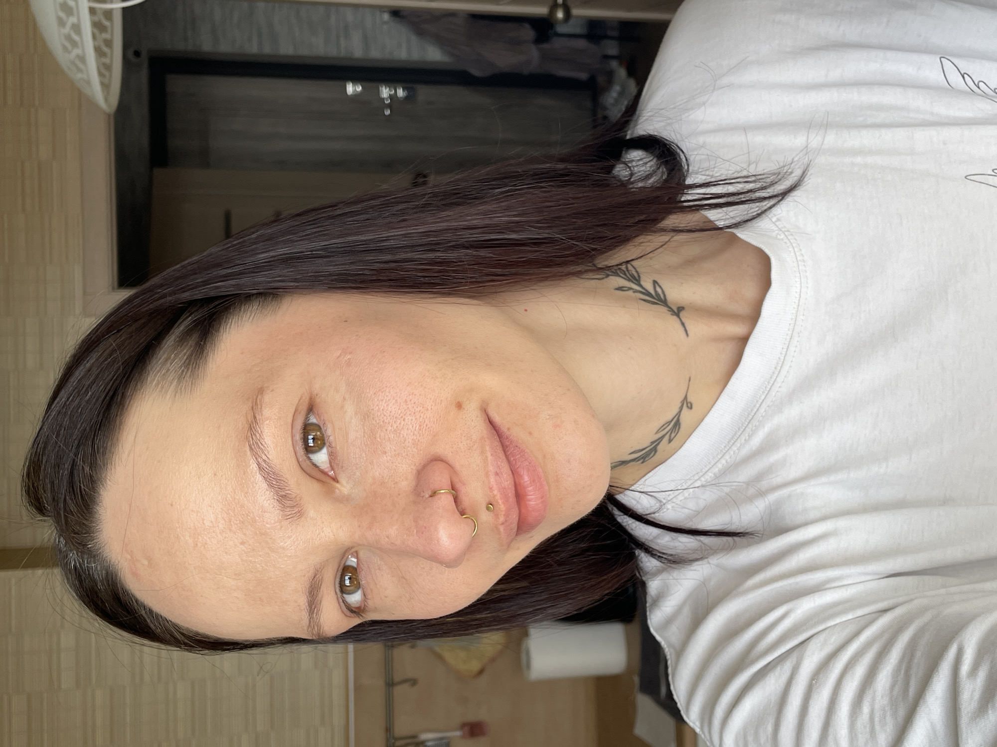 A woman is centered in the image, wearing a white t-shirt. She has dark hair and visible tattoos on her neck. She also has two visible piercings on her nose. The background includes a door, a fan, and what appears to be a kitchen counter with a paper towel roll. No information is available from the image to confirm or deny that the photo was taken in Izhevsk, Russia.