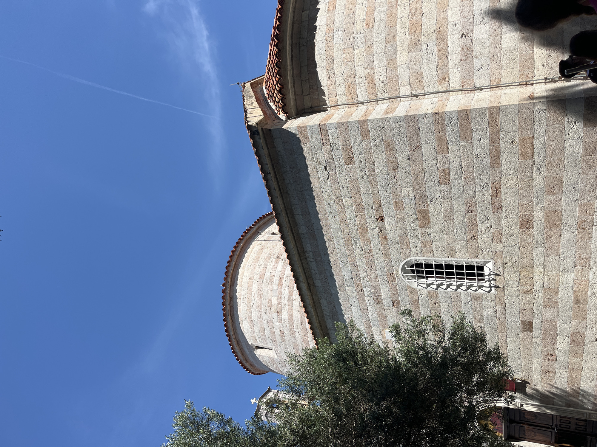 A low-angle, rotated view depicts an exterior stone building with a curved facade and red-tiled roof, positioned against a clear blue sky with a contrail. The structure's facade features vertical bands of light and dark stone. A small, barred window punctuates the wall, partially obscured by green foliage of a tree in the foreground. The building is the Church of Santa Maria in Punta, located in the city of Budva, Montenegro. Parts of two people are visible in the upper right corner.