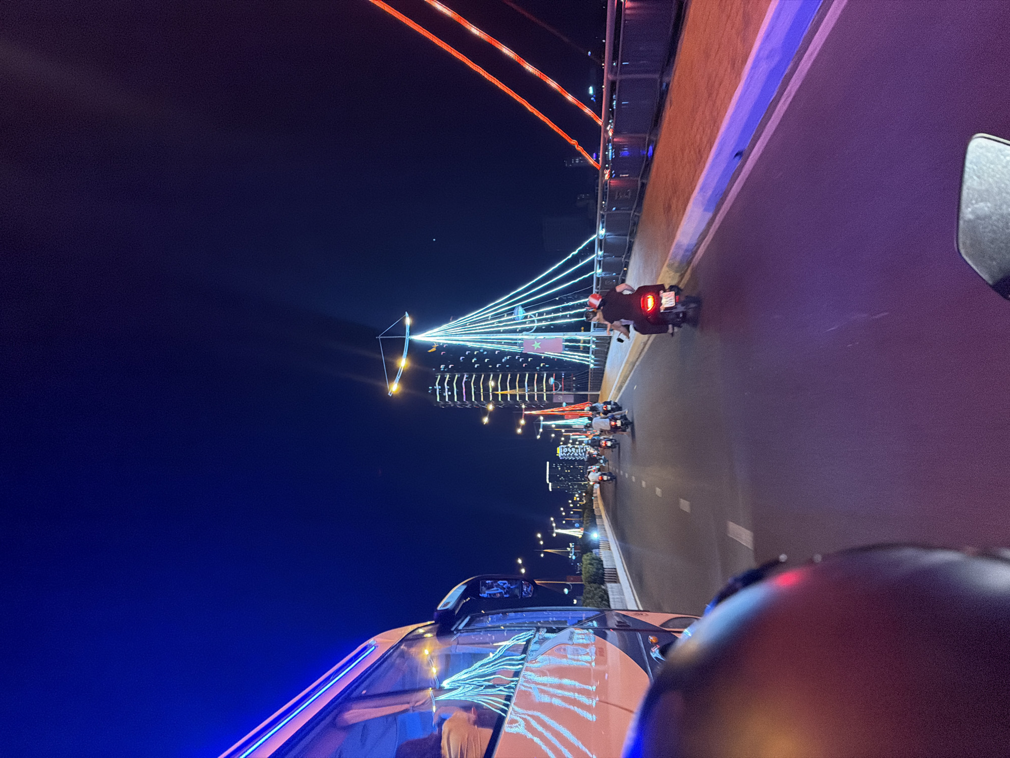 The image depicts a nighttime scene on a road in Nha Trang, Vietnam. A bridge with illuminated white cables extending from a central pylon structure is visible. The bridge also features red lighting along its upper structure. Several motorbikes are traveling on the road. The image is taken from the perspective of someone in a vehicle, with part of the vehicle's hood and rearview mirror visible in the foreground. A dark helmet occupies part of the lower right. The sky is dark. Buildings with lights are visible in the distance, lining the road. The road surface is lit by streetlights.