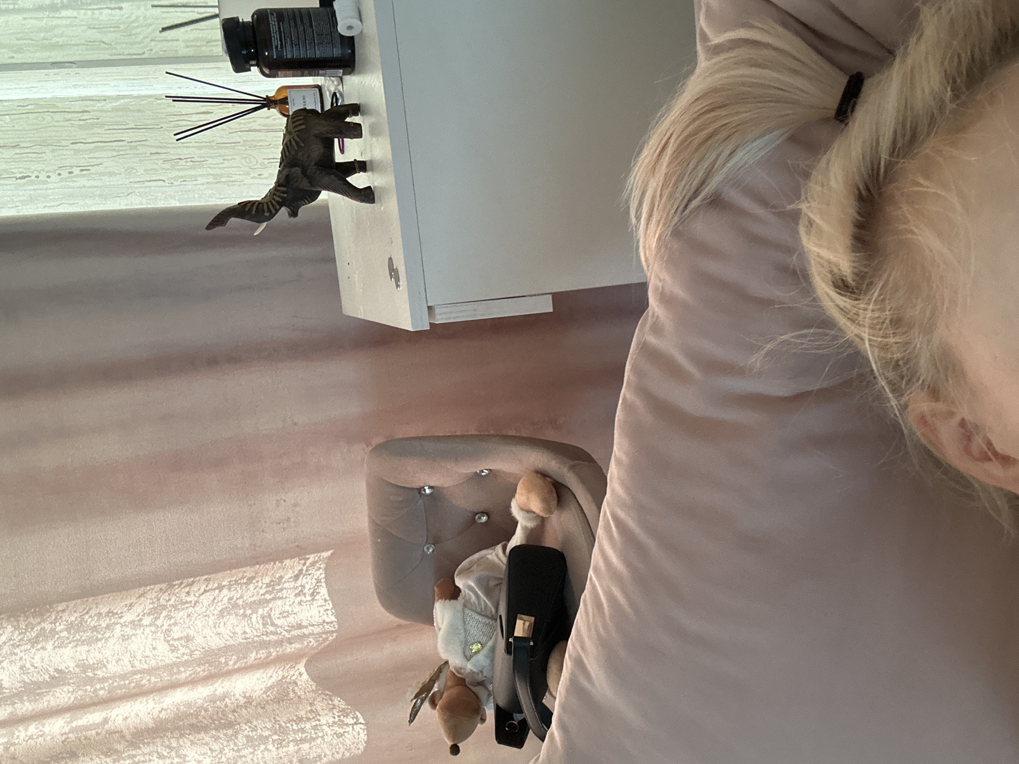 A close-up shot, potentially from Высокий, Russia, presents a bedside scene. Part of a person with blonde hair is visible on the right side of the frame. Next to the person is a light pink bed covering and a bedside table in the top center. The table has a diffuser, a bottle, and a toy dinosaur on top. Underneath the table, on the left of the shot, there's a small grey padded stool with a plush toy and a black bag. The background is a wall with a patterned wallpaper.