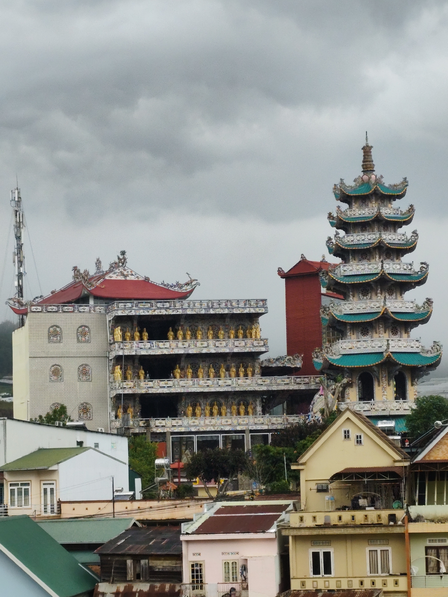 The image shows a cityscape of Xuân Thành, Vietnam, under a cloudy sky. A tall, multi-tiered pagoda with a green and white roof stands prominently on the right side of the image. Adjacent to it is a building with a red roof, featuring multiple rows of golden statues. In the foreground, various residential buildings with different colored roofs, including green, brown, and pink, are visible. A telecommunications tower is visible to the left of the buildings, behind the pagoda.