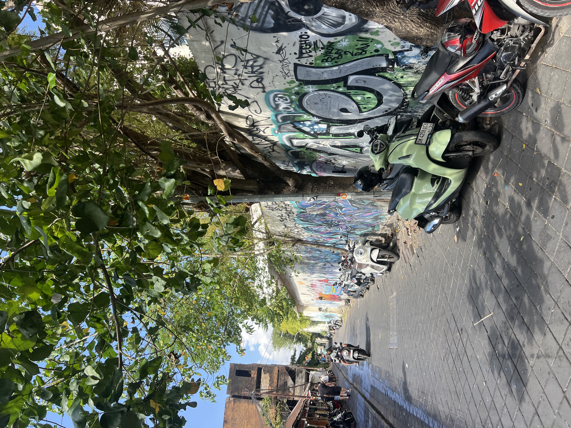 In Kuta, Indonesia, a narrow, paved street runs between buildings, lined with parked scooters and motorcycles. The street is illuminated by dappled sunlight filtering through the branches of a large tree. On the right side of the street, a concrete wall is covered in vibrant graffiti, featuring large, bold lettering and abstract designs in green, black, and white. A light green scooter is parked in front of the graffiti wall, followed by a red and black motorcycle. In the distance, more scooters are parked along the left side of the street, next to shops with awnings. A few people are walking on the street, and one person is riding a scooter. The sky above is a clear blue, with a few wispy clouds.