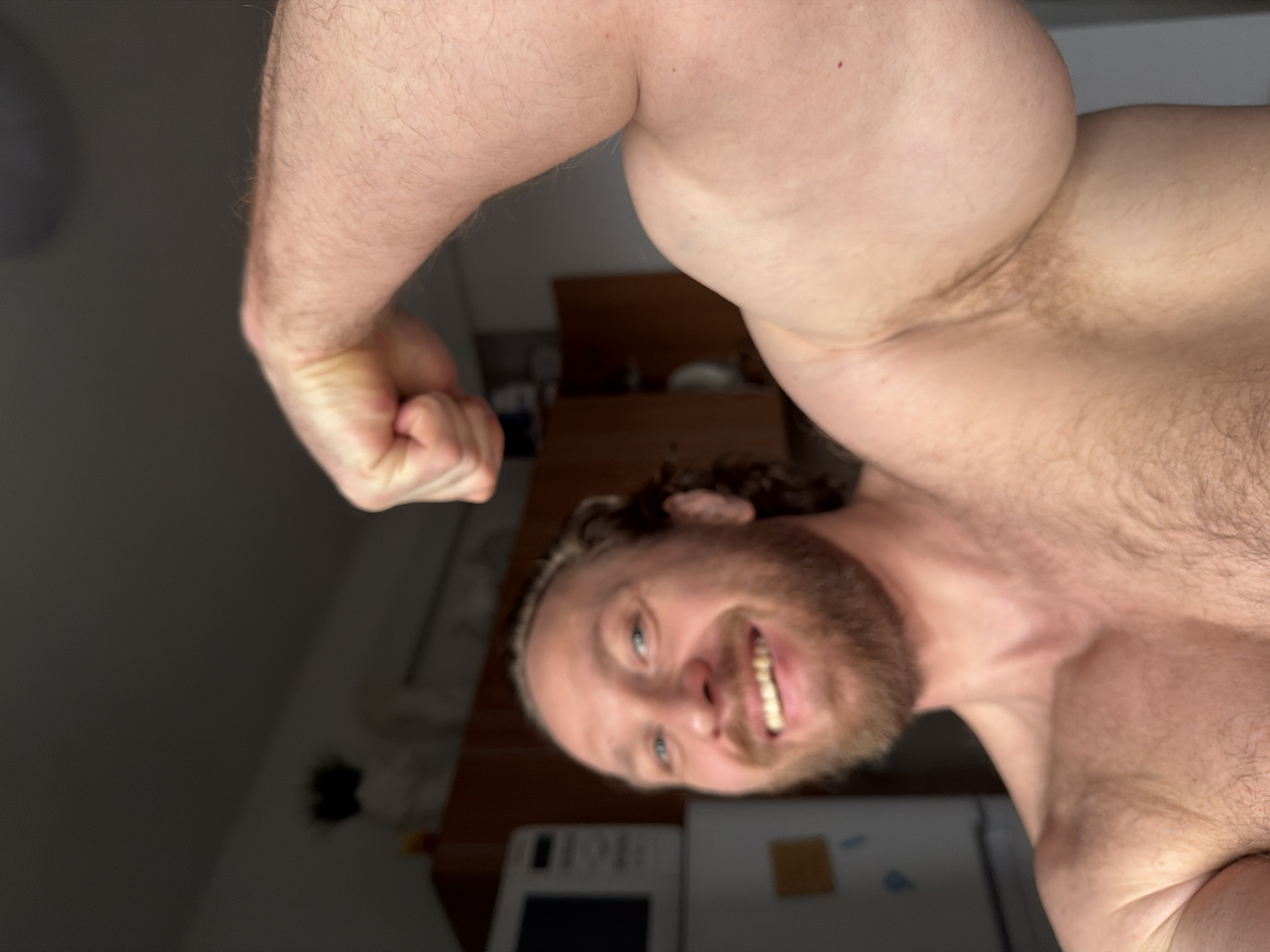 A shirtless man with a beard is posing with his arm flexed, displaying his bicep muscle. He is smiling and looking directly at the camera. The background appears to be a kitchen or dining area, with a microwave and wooden furniture visible. The photograph may have been taken indoors, and its location cannot be verified as Saint Petersburg, Russia.