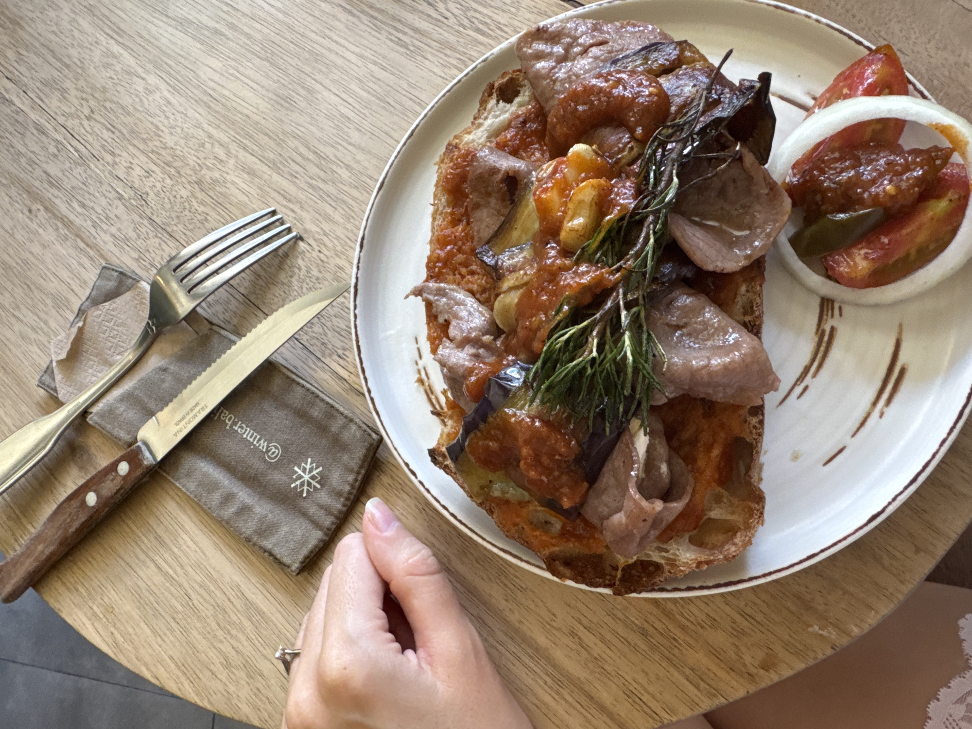 The image depicts a table setting with a plate of food, cutlery, and a hand. The food on the plate appears to be a slice of bread topped with meat, vegetables, and a sprig of rosemary. A small side of sliced tomatoes and onions with a relish is also present. A fork and knife rest on a small cloth napkin to the left of the plate. A hand is partially visible in the lower left corner of the image. The table has a light wood grain finish. The location of the scene is Unknown, Unknown.