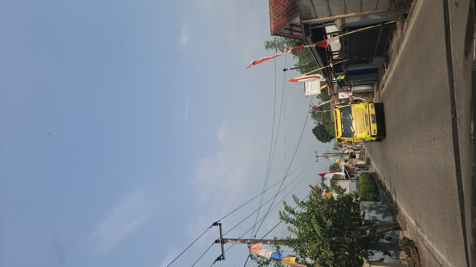 A yellow truck is driving down a paved road in Sirnagalih, Indonesia. The road is lined with houses and trees. There are several flags displayed, indicating a national celebration. The sky is blue with some clouds. The image appears to be taken from a vehicle.