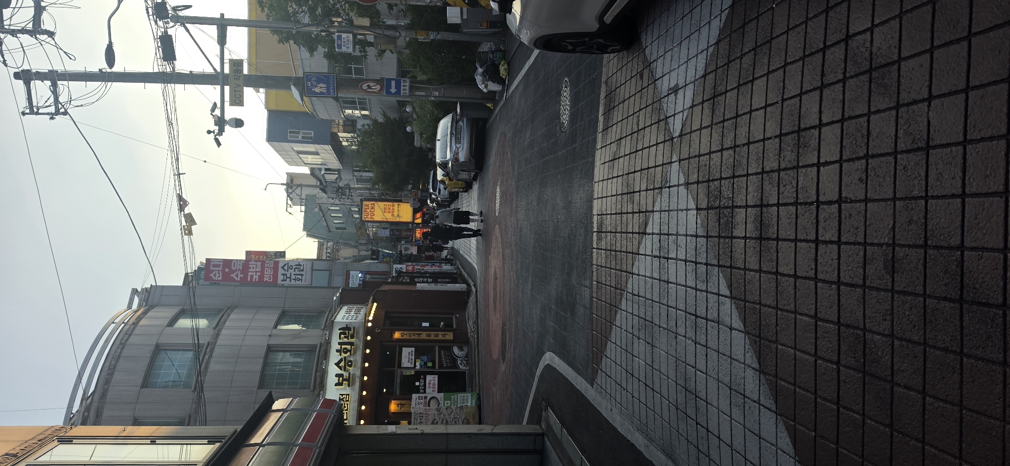 This is a street-level, medium-angle, vertical shot of a street in Cheongju-si, South Korea, during the golden hour. The street is paved with dark asphalt, with a section of patterned tiles in the foreground. On the left, a building with a curved facade and large windows is visible, with a red and white awning. Next to it is a shop with illuminated signage and a menu board. Across the street, a row of businesses with various signs and awnings stretches into the distance. A few cars are parked along the right side of the street. Two individuals, a man and a woman, are walking away from the camera down the street. Overhead, a complex network of electrical wires and utility poles crisscrosses the sky, with traffic signs and a CCTV camera visible on one of the poles. The sky is a soft yellow and orange, indicating either sunrise or sunset. The overall atmosphere is that of a quiet, ordinary street scene in an urban environment.
