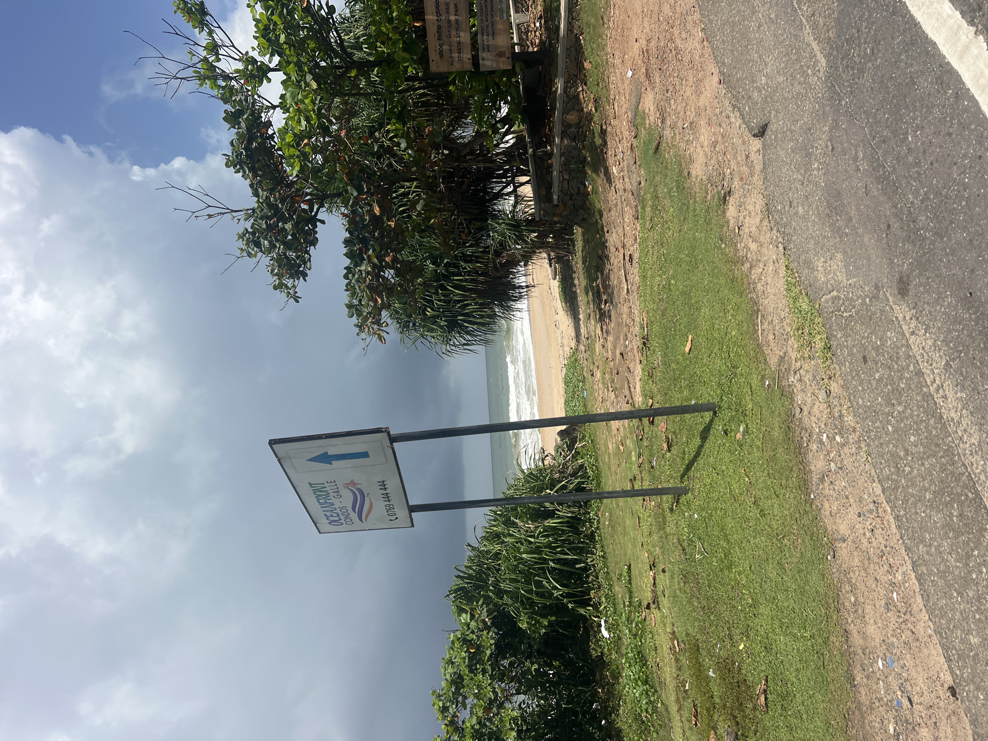 The image depicts a beachfront scene in Galle, Sri Lanka. A white sign with blue text and an upward-pointing blue arrow is visible on the left, indicating directions for "OCEANFRONT CONDOS - GALLE" and a phone number "0769 444 444". To the right of the sign, a sandy beach meets the ocean with gentle waves. Lush green vegetation, including palm-like plants and trees, borders the beach and the roadside. A wooden sign with text in a foreign language, possibly Sinhala, is partially visible behind the trees on the right, along with English text that includes "COASTAL LEISURE PARK" and "DEPARTMENT OF COAST AND COASTAL RESOURCES MANAGEMENT". The sky is a mix of blue with white clouds and darker, more ominous clouds on the left. The foreground shows a paved road and a grassy area with scattered leaves and debris. No people are present in the scene.