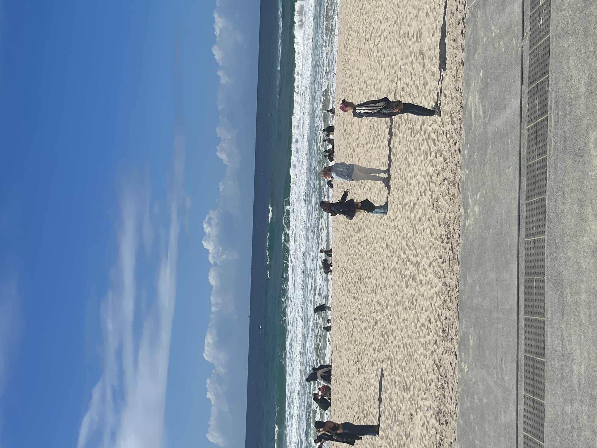 The image depicts a bright, sunny day at a beach, likely in the city of Gold Coast, Australia, given the context of the provided crops. The scene is divided into three main horizontal sections: the sky, the ocean, and the beach.
The sky is a vibrant, clear blue with scattered, fluffy white clouds, suggesting fair weather. The ocean is a deep turquoise with white-capped waves rolling towards the shore, indicating moderate surf. A faint silhouette of a distant boat can be seen on the horizon.
The foreground features a sandy beach populated by a number of people. In the middle ground, there are several individuals and small groups scattered across the sand. To the left, a woman in a dark jacket and jeans stands facing away from the camera. Further back, a group of people are seated or lying on the sand near the water's edge. In the center of the beach, two women are talking, one wearing a light blue hooded sweatshirt and the other a dark jacket. To their right, a person in a dark jacket and a red cap stands facing the ocean. Other individuals are dispersed across the beach, some walking and some seemingly observing the ocean.
The immediate foreground shows a concrete pathway with a metal grate, typical of a beachfront promenade or walkway. The sand itself appears well-trodden, with footprints visible. The overall impression is of a lively, public beach on a pleasant day.