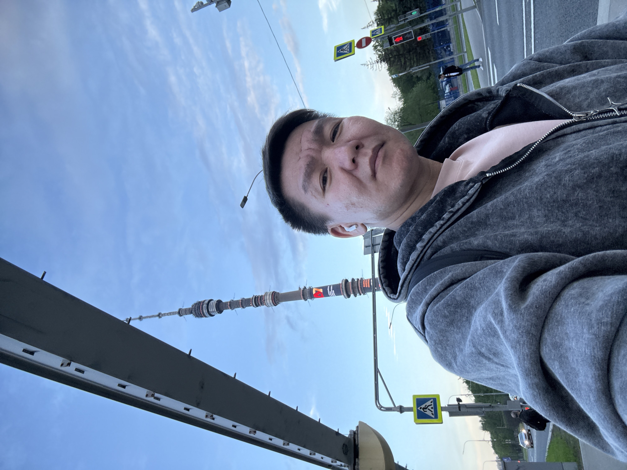 A middle-aged Asian man in a gray zip-up hoodie is taking a selfie on a bridge. Visible behind him is the Moscow TV Tower, suggesting the location is in or near Moscow, Russia. Further details include traffic lights, pedestrian crossing signs, and a road with cars. The sky is visible in the background and appears partly cloudy. The perspective is tilted, presenting an off-center view.