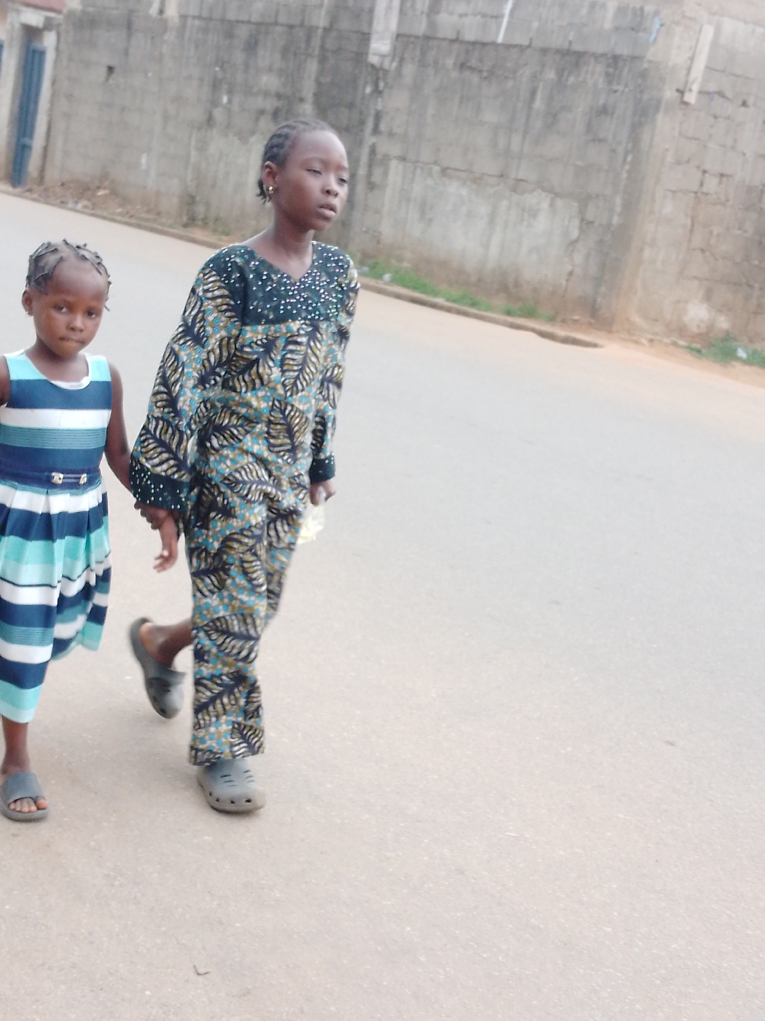 Two young girls are walking together on a paved road. The younger girl is on the left, wearing a blue and white striped dress with a dark blue belt, and gray sandals. She is holding the hand of the taller girl, who is wearing a printed long-sleeved top and matching pants, with gray closed-toe sandals. Behind them is a concrete wall, partially covered in darker patches and some small patches of green vegetation. The location appears to be a residential street. The city and country are not discernable from the image.