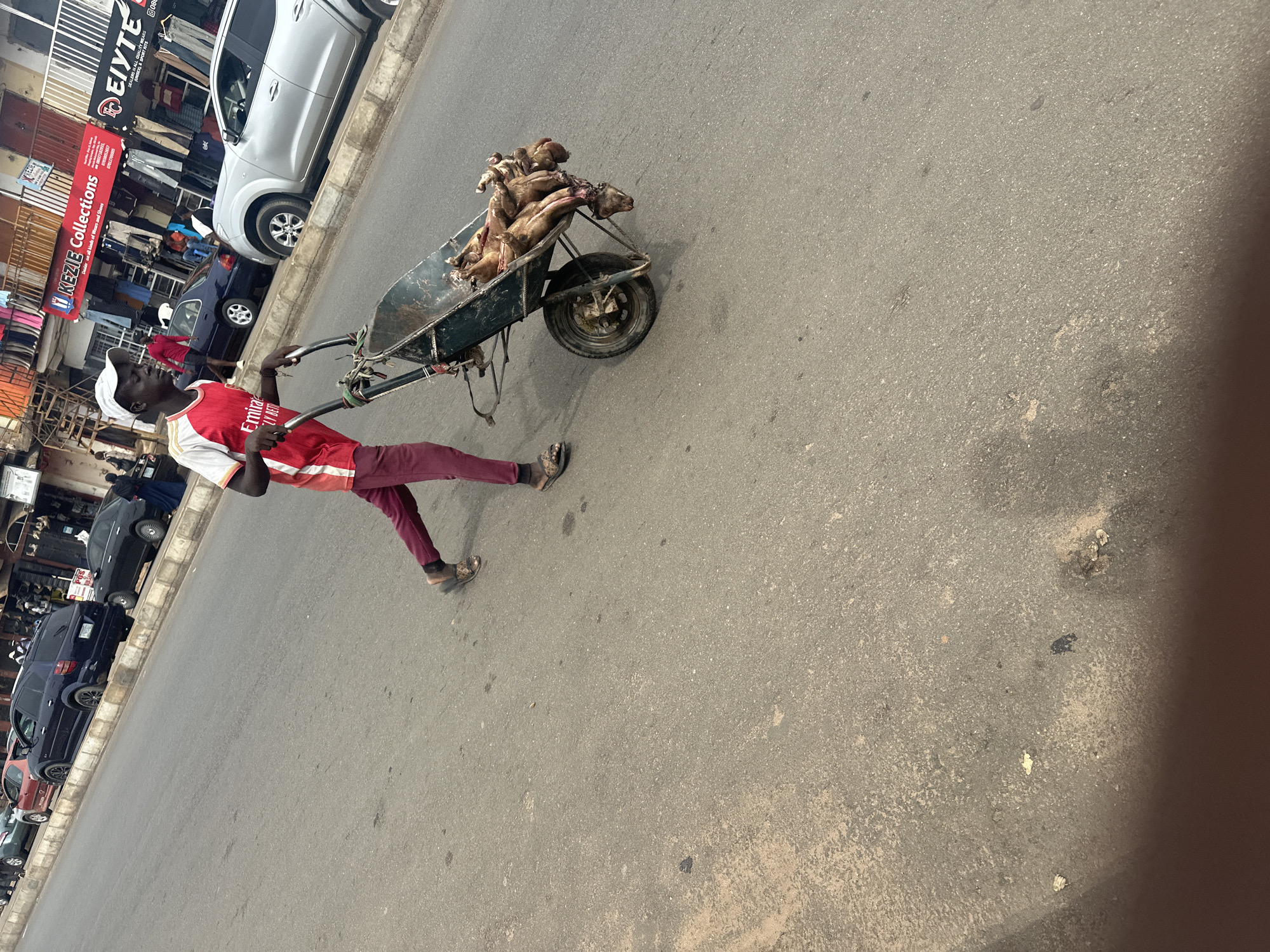 A young boy pushes a wheelbarrow filled with several dressed goats down a street in Jos, Nigeria. The boy, wearing a red and white shirt and maroon pants, is walking on the asphalt road. The wheelbarrow is dark green with a single wheel in the front. Several shops are visible in the background along the street, with one displaying a sign that reads "KEZIE Collections." The sky is overcast, and the overall lighting suggests it is daytime. Other vehicles, including cars and a motorcycle, are also present on the street.