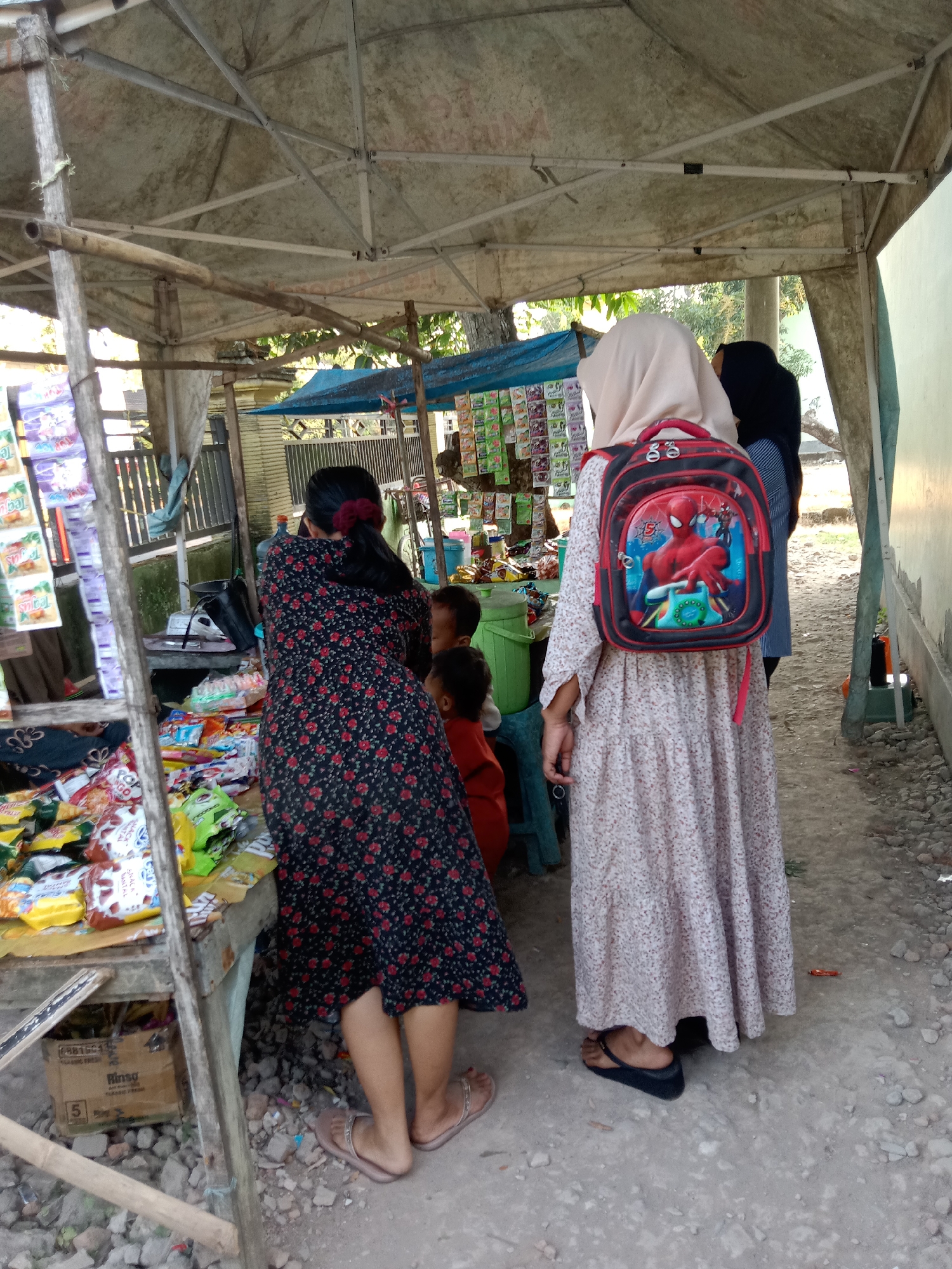 In Sugihwaras, Indonesia, a street vendor's stall displays an assortment of packaged snacks and goods. Two women and two children are present. One woman, wearing a floral print dress, is facing away from the camera, examining the merchandise. The other woman, also facing away, wears a light-colored hijab and a long, patterned dress, and carries a Spider-Man themed backpack. Two children are partially visible near the stall, one wearing an orange shirt. The stall itself is covered by a tarp and constructed with bamboo and wood. The ground is composed of dirt and small stones.