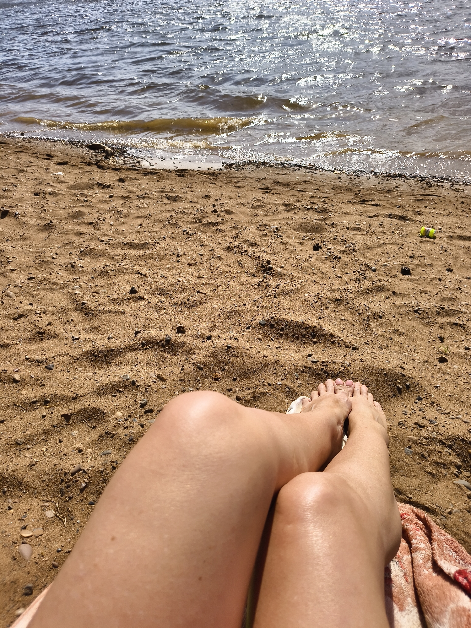 A person is lying on a sandy beach near the water, with their legs visible in the foreground. The sand is a mix of brown and tan, with small pebbles scattered throughout. In the background, the water is a dark color, with sunlight glinting off the surface. A yellow object sits in the sand, behind the person's legs. The location is Unknown, Unknown.
