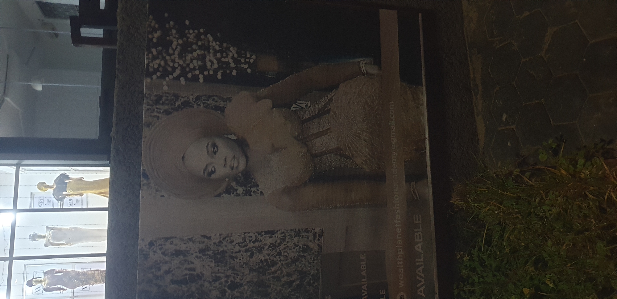 A billboard features a glamorous woman in traditional attire with a large headwrap and ornate dress. The billboard is set against a marbled background with sparkling embellishments on the right. The text on the billboard reads "AVAILABLE" and "wealthplanetfashionacademy@gmail.com". In the background, a store window displays mannequins dressed in elaborate gowns, suggesting the location is a fashion house or academy. The store window is framed by what appears to be a building with white walls. Below the billboard, the ground is paved with dark hexagonal tiles and a patch of lush green grass and weeds. The overall scene depicts a commercial advertisement in an urban setting, likely at night, given the lighting. The location is identified as Narayi, Nigeria.